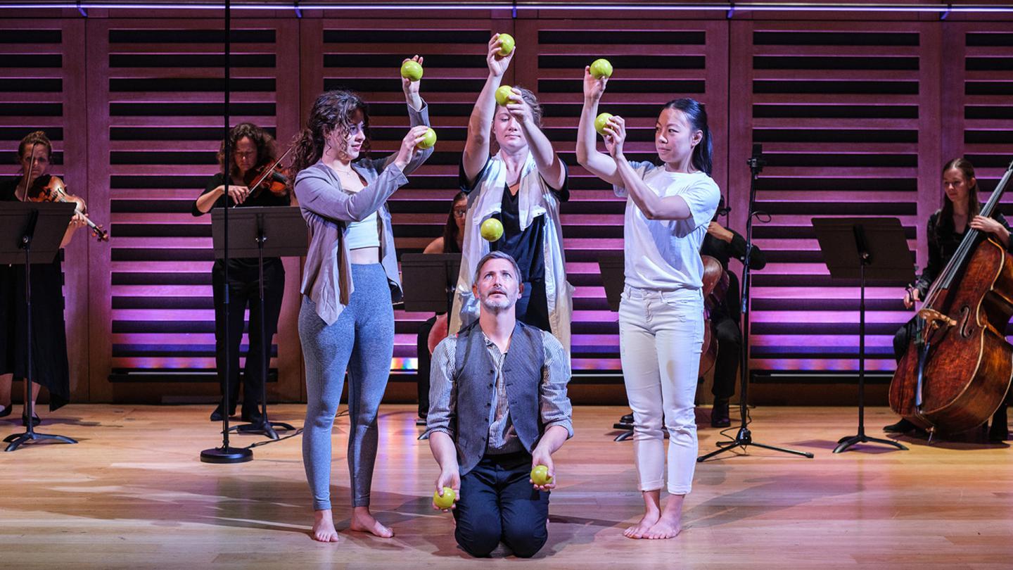 One man juggles while kneeling on the floor while three women stand around him passing and catching balls