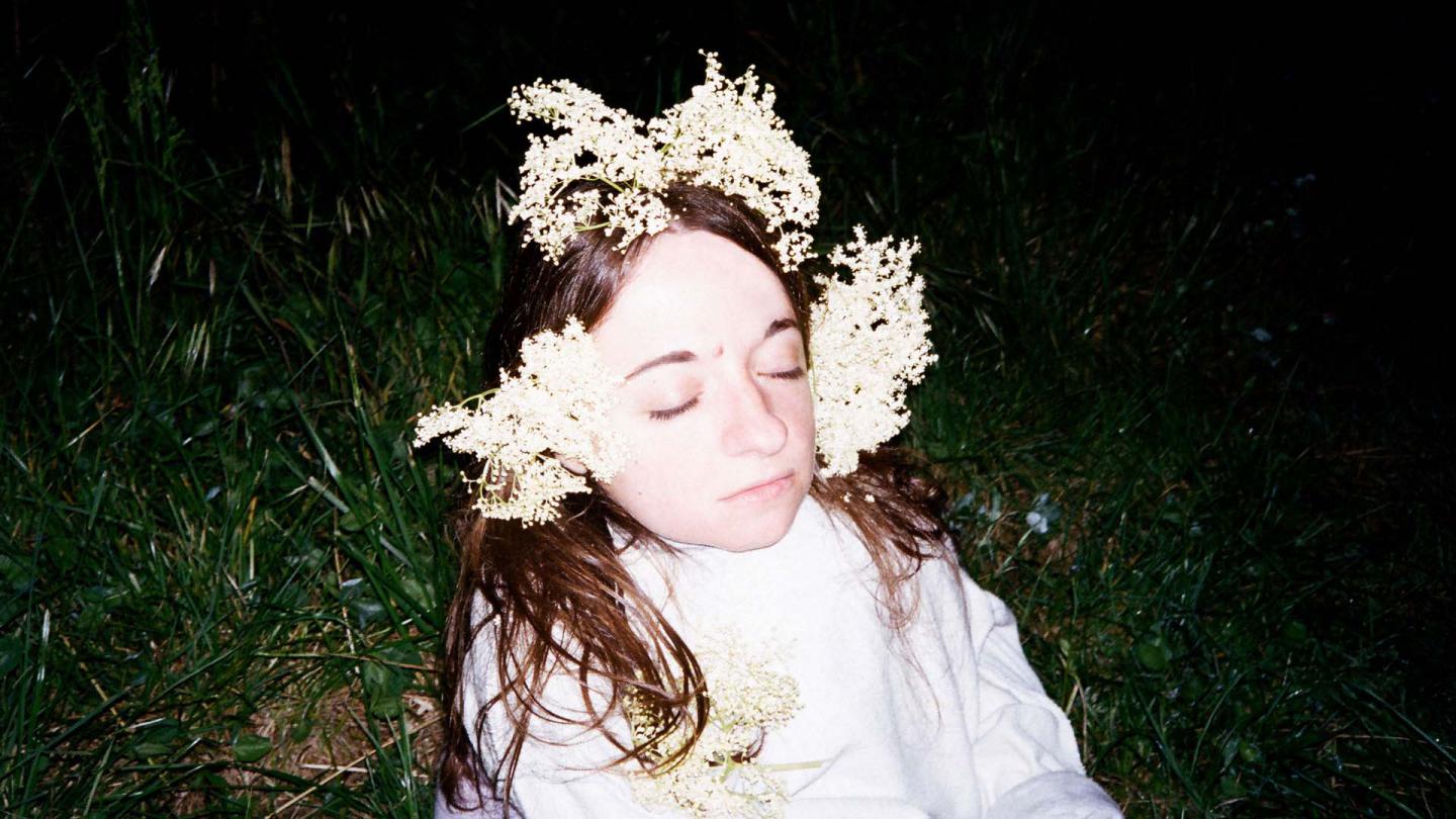 PErson on grass in the dark with her eyes closed, lit brightly with white flowers in her hair and a wearing white shirt