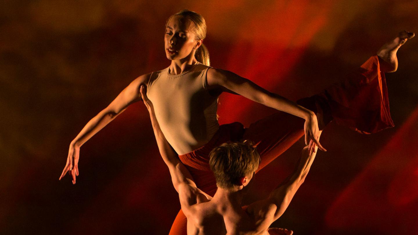 Bathed in red light and male dancer holds upa female dancer as she holds a pose with her arms straight and her fingers pointed down