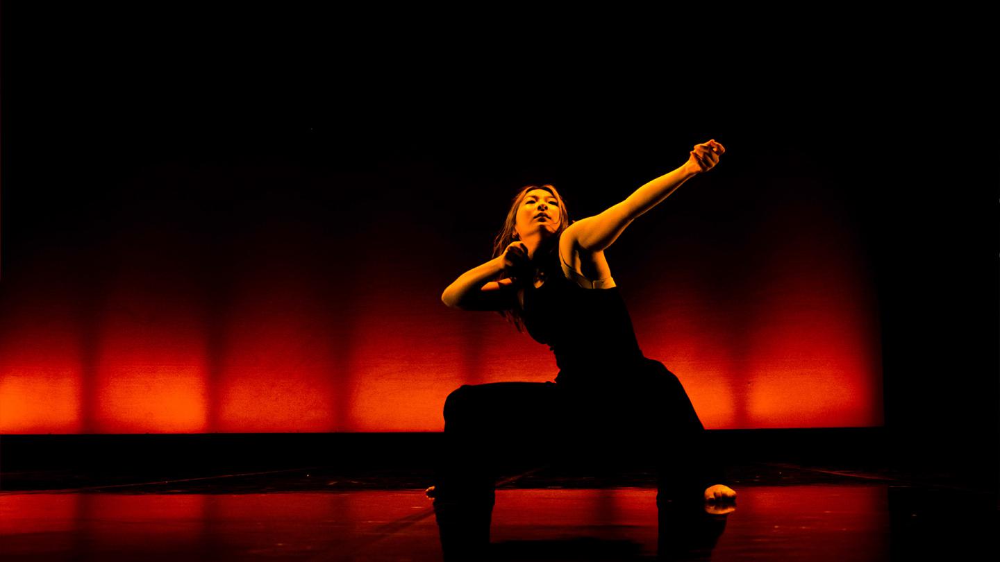 Dancer wearing black trousers and a black vest squat, one arm bent and the other extended as drawing an archery bow.