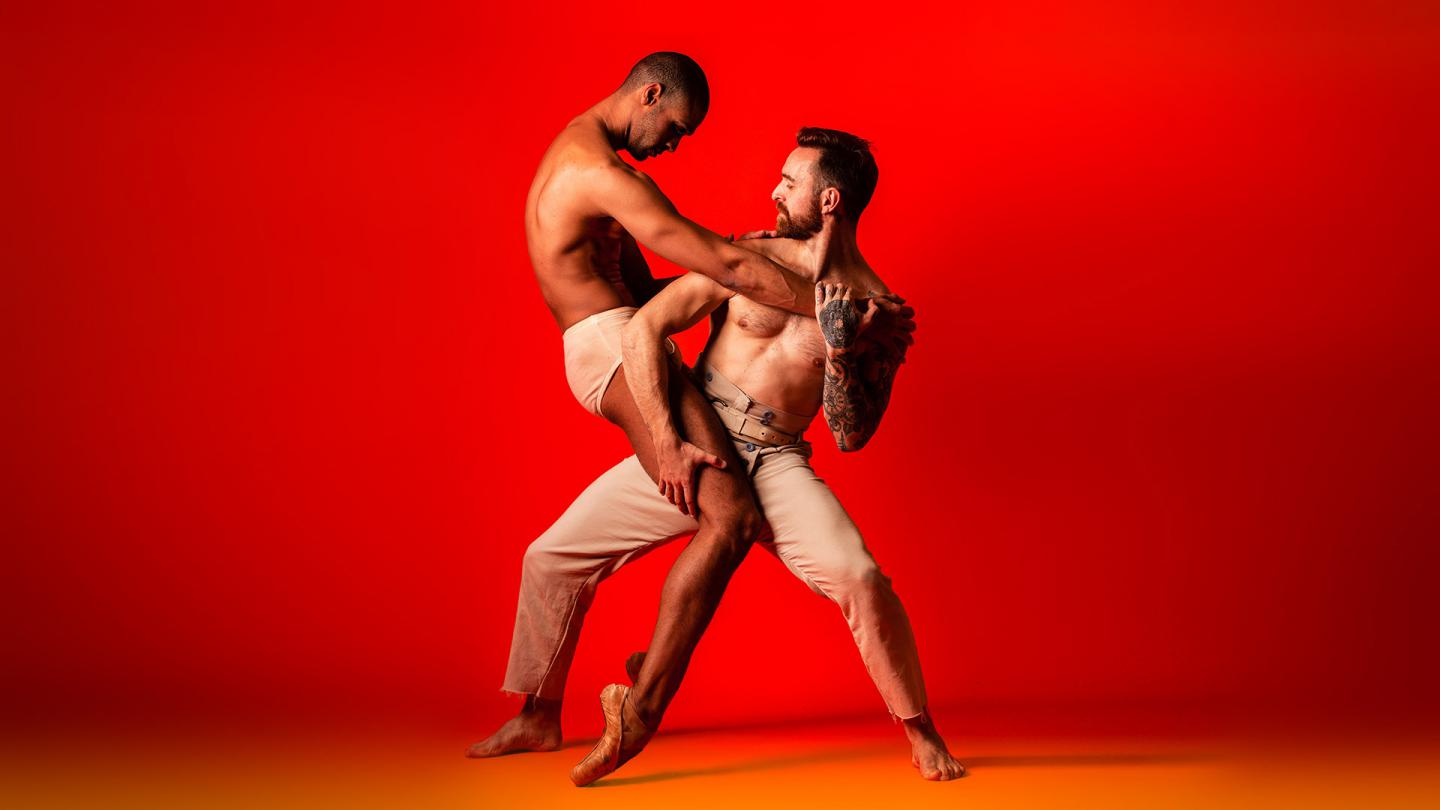 Against a red backdrop, two men dance, embracing each other, simultaneously leaning in an away, wearing pale beige