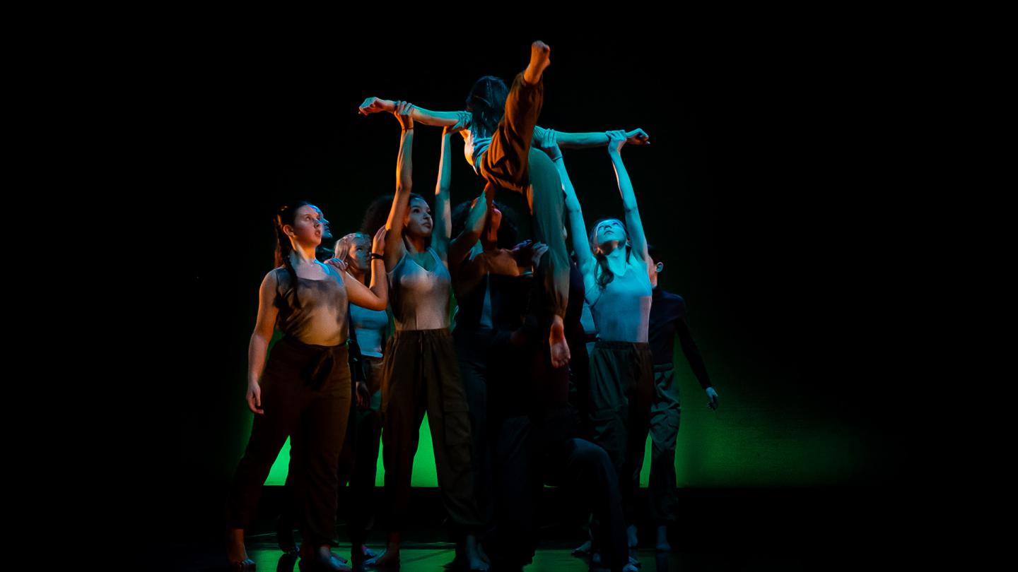 Against a black backdrop, many dancers hold up one with their arms apart, one leg arabesque and the other pointing to the ground