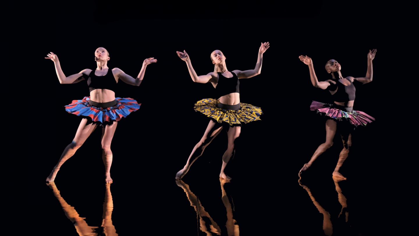 Against a back background and shiny black floor, three dancers dressed in colourful tutus pose with their arms raised, bent at the elbow