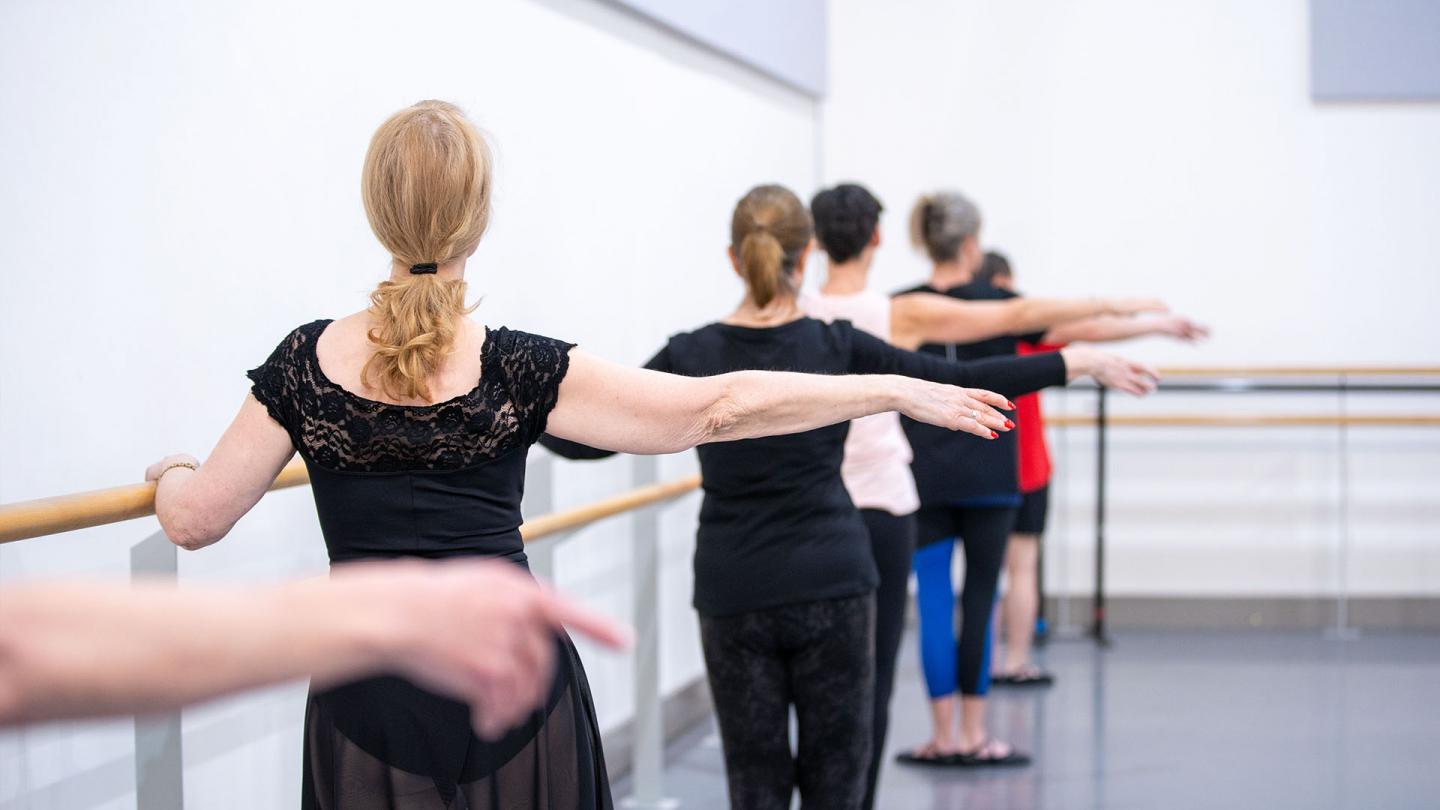 A row of dancers at the barre, one hand held out level with their shoulders, the other hand holding the barre, their ankles together in first