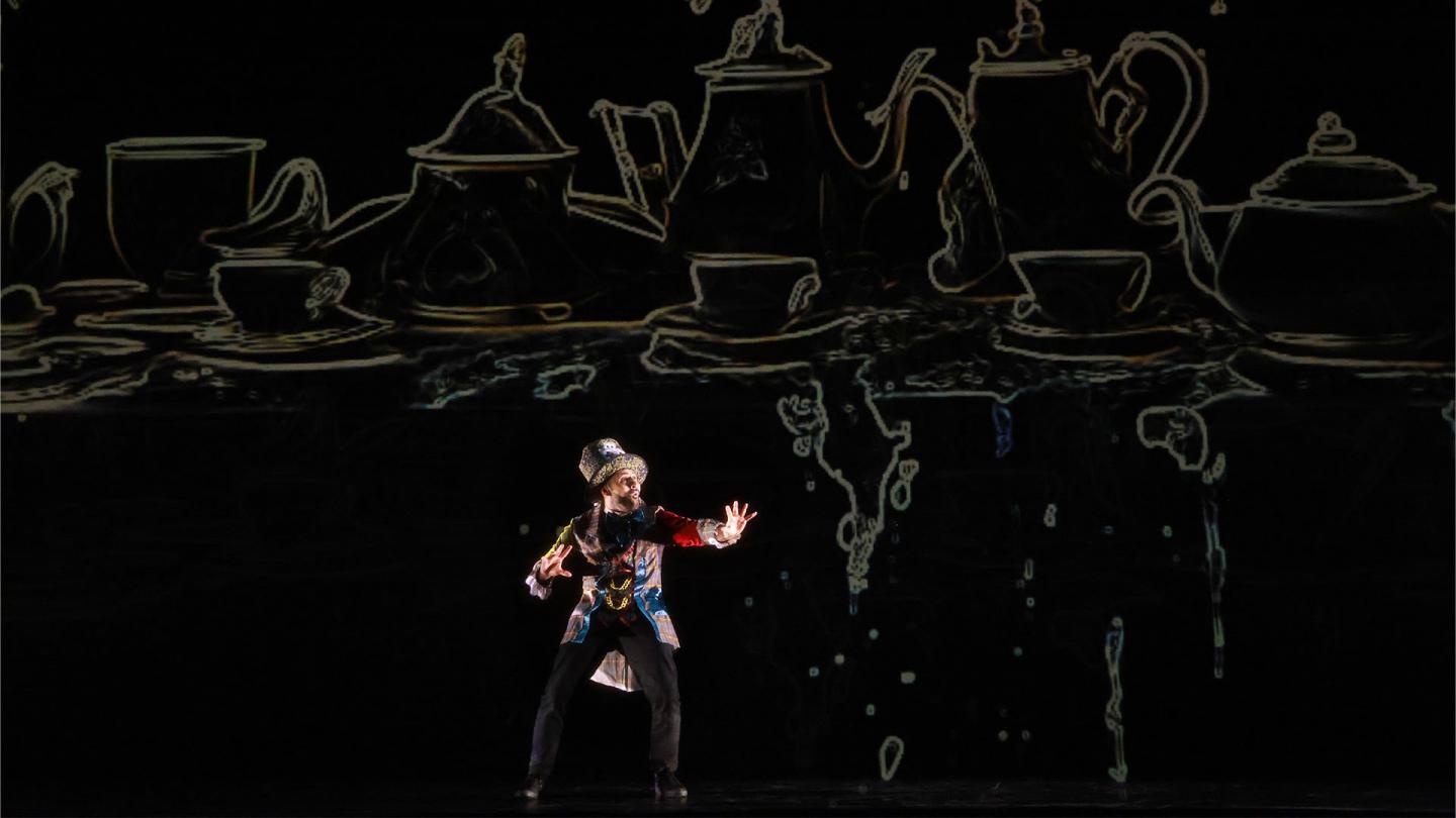 A man in a large top hat motions in front of giant, stylised crockery