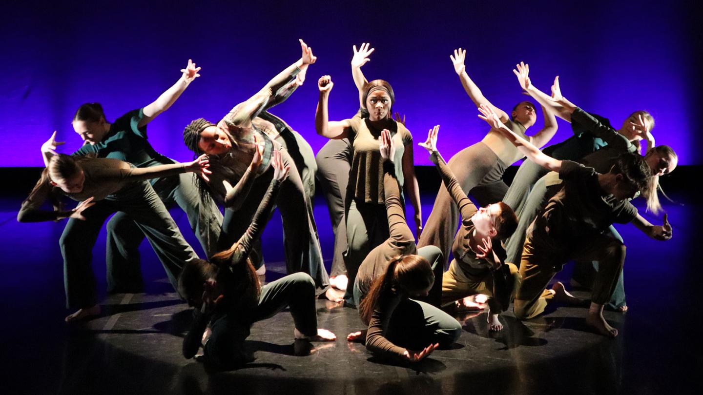 Against a blue and purple backdrop, a figure stands with one arm raised in a defiant gesture surrounded by dancers shielding themselves from her brilliance.