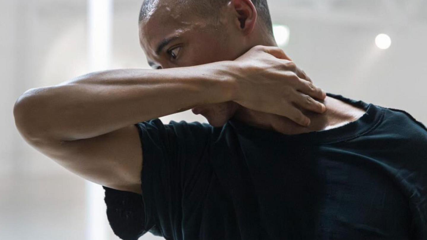 A dancer in a black shirt covers their face with their forearm while holding their hand to the side of their neck.