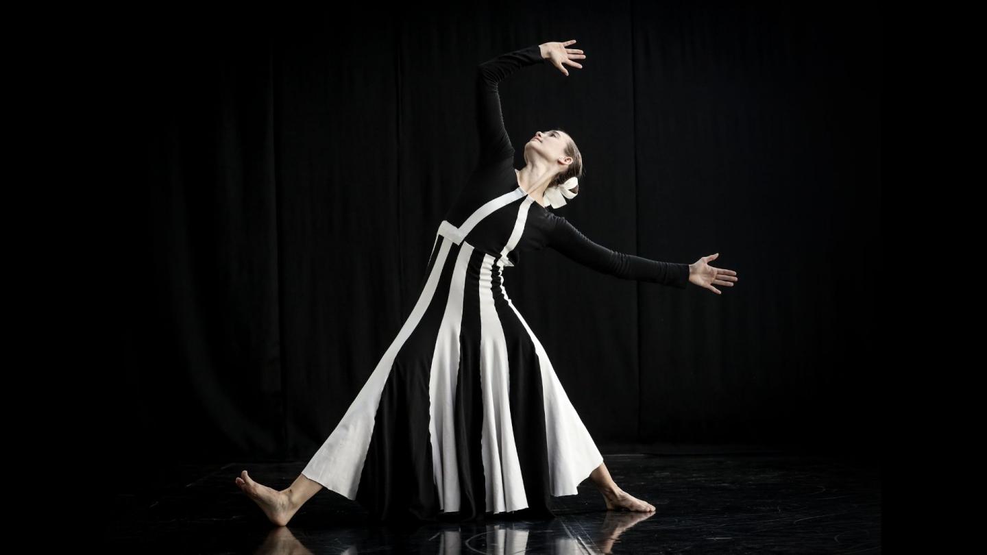 On a black background, a dancer in a black and white striped dress poses with an arm held above their head and their legs spread in a wide stance.