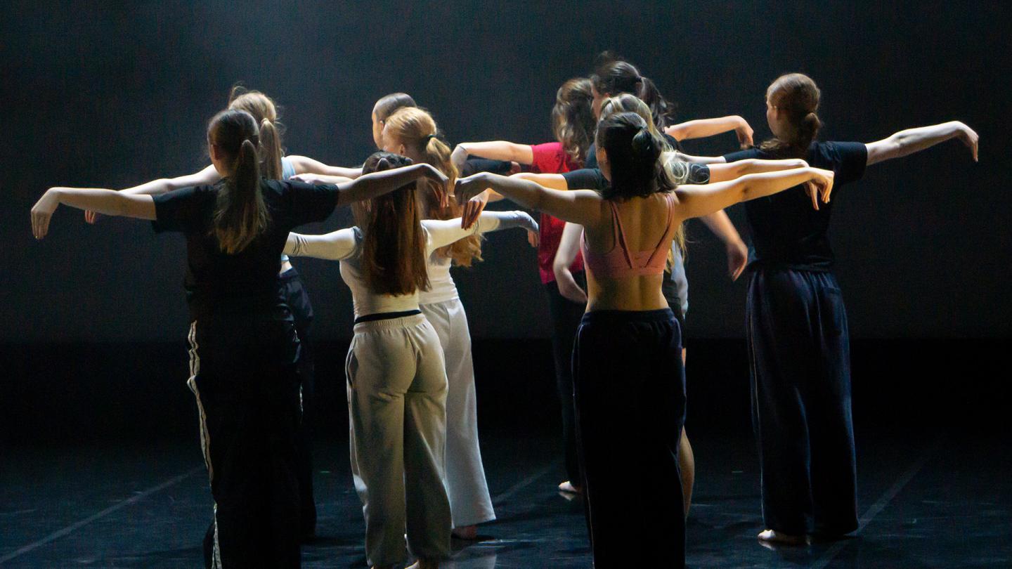 Troop of dancers turned away from the viewer, each standing with their arms straight out level with their shoulders
