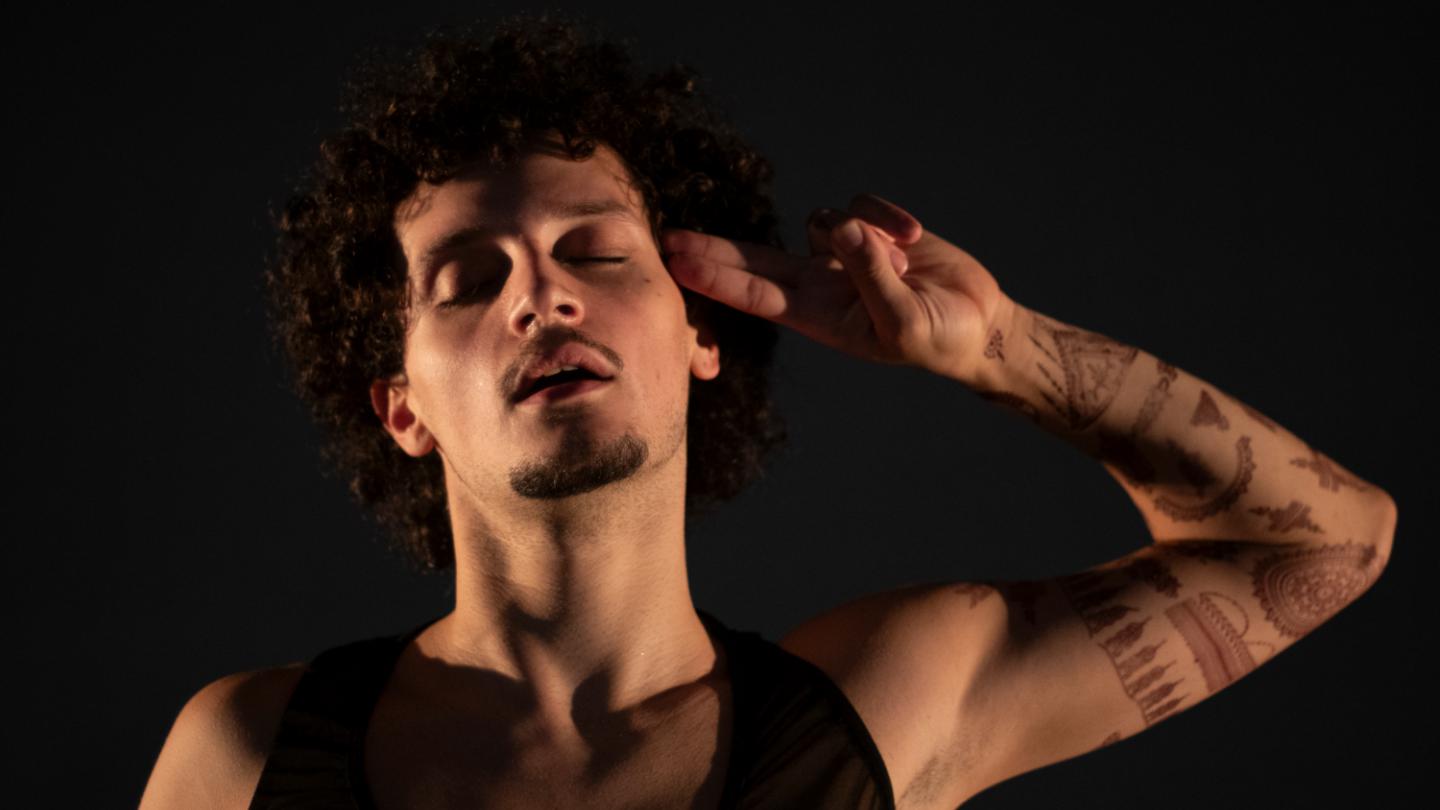 On a black background, a dancer faces the camera with their eyes closed and holds two fingers to the side of their head.