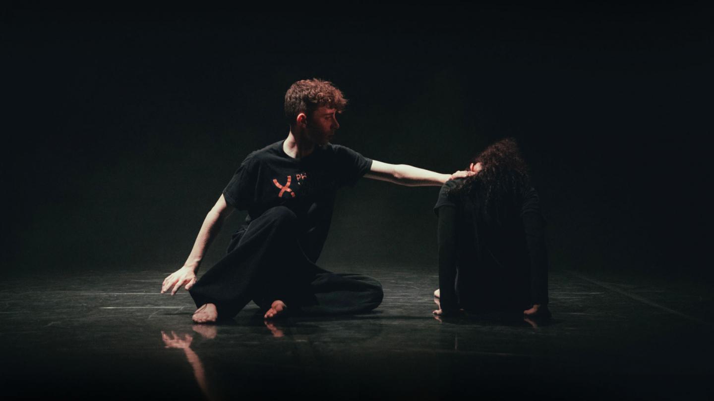 Dancers on the dark floor , lit sparingly from above, one reaching out to the other who has their back to us and head down