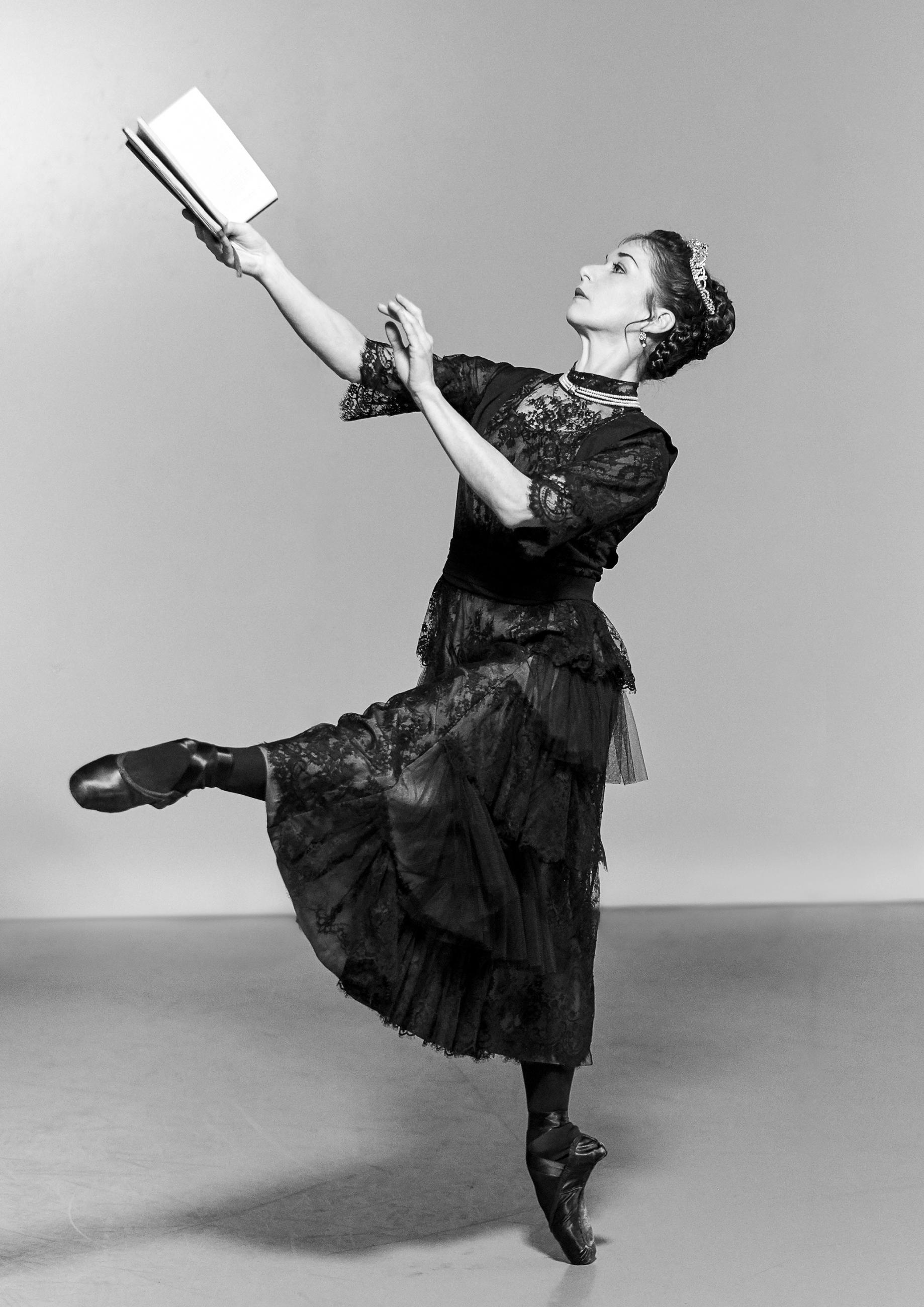 Pippa Moore dancing as Older Beatrice at the Victoria Photoshoot. She is holding a book in her outstretched arm. The image is black and white, she is wearing a dark lace covered dress.