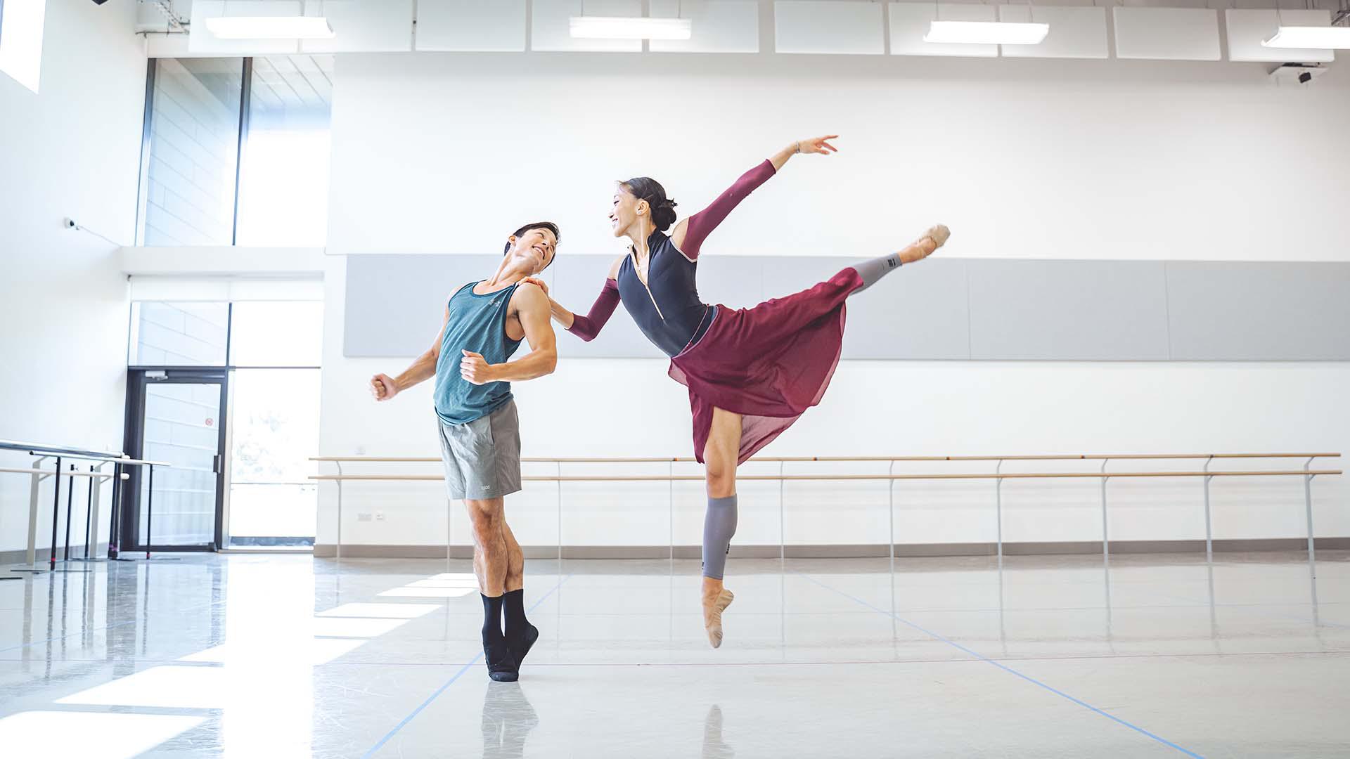 A male dancer stands on his toes while a female dancer is landing a full jete while holding his shoulder