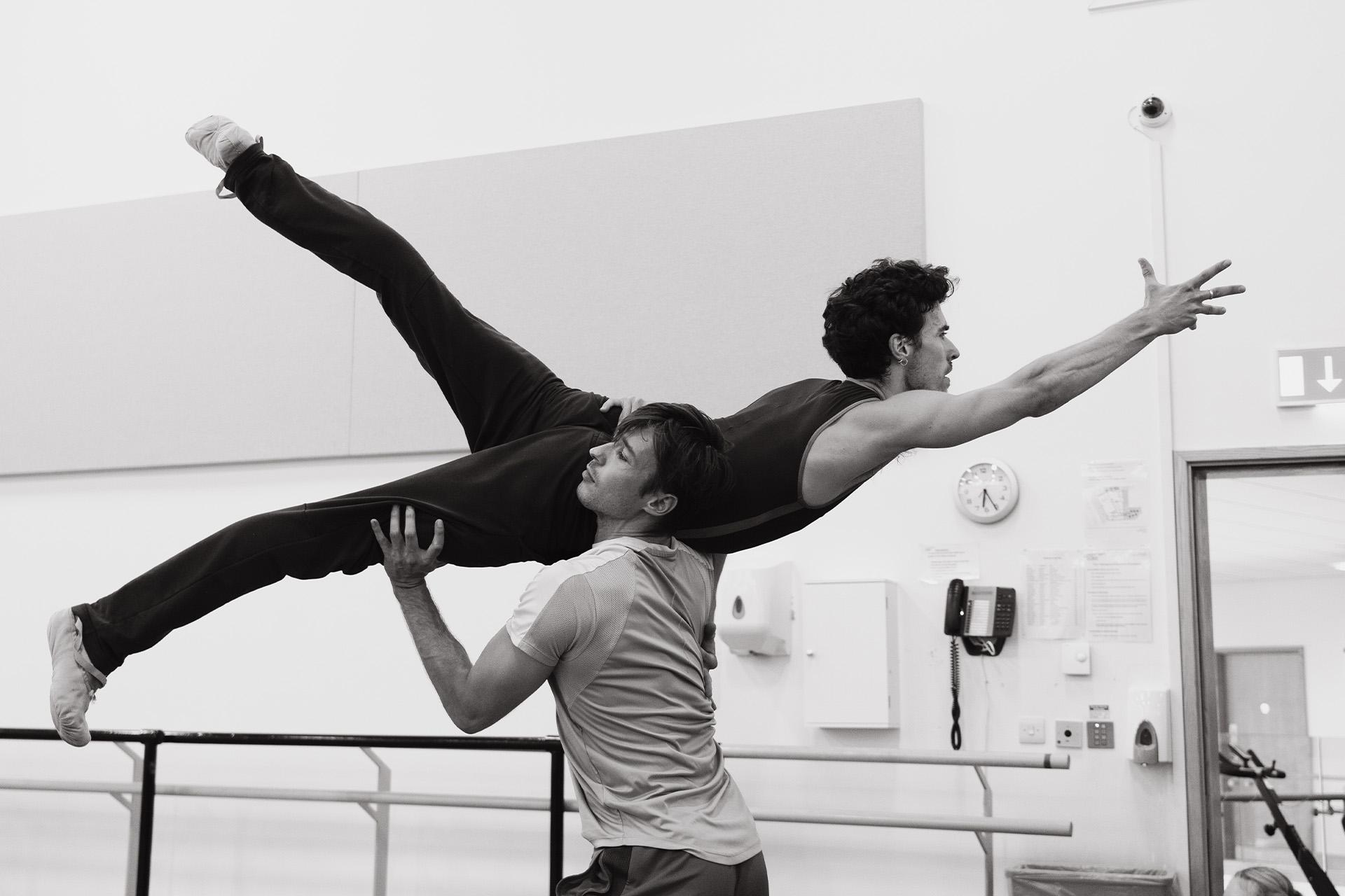 Black and white photo of two male dancers, one holding the other on his shoulder while the others arms and legs a stretched out