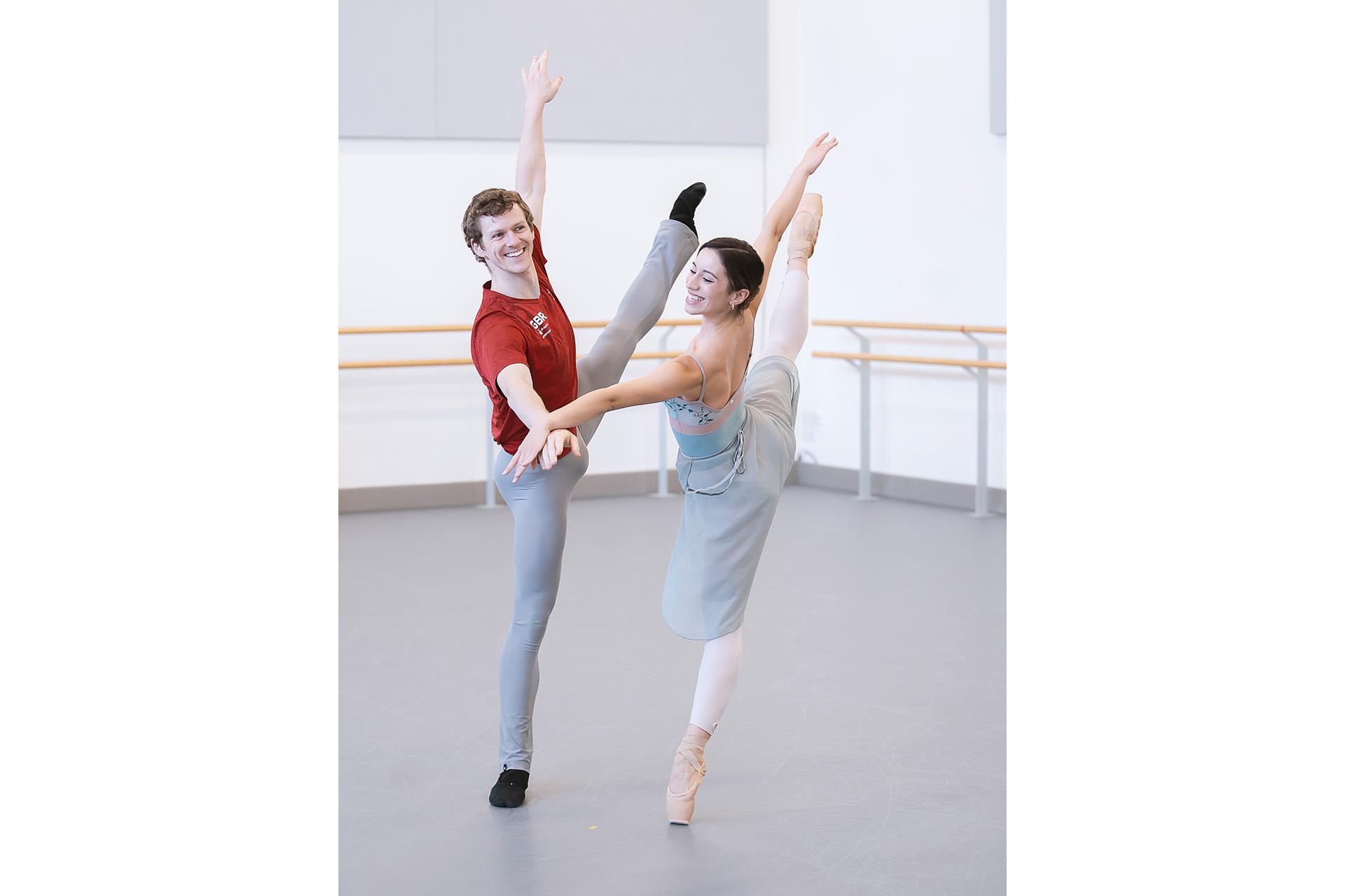 Two dancers hold hand while each are on one foot, the other raised to shoulder level while smiling