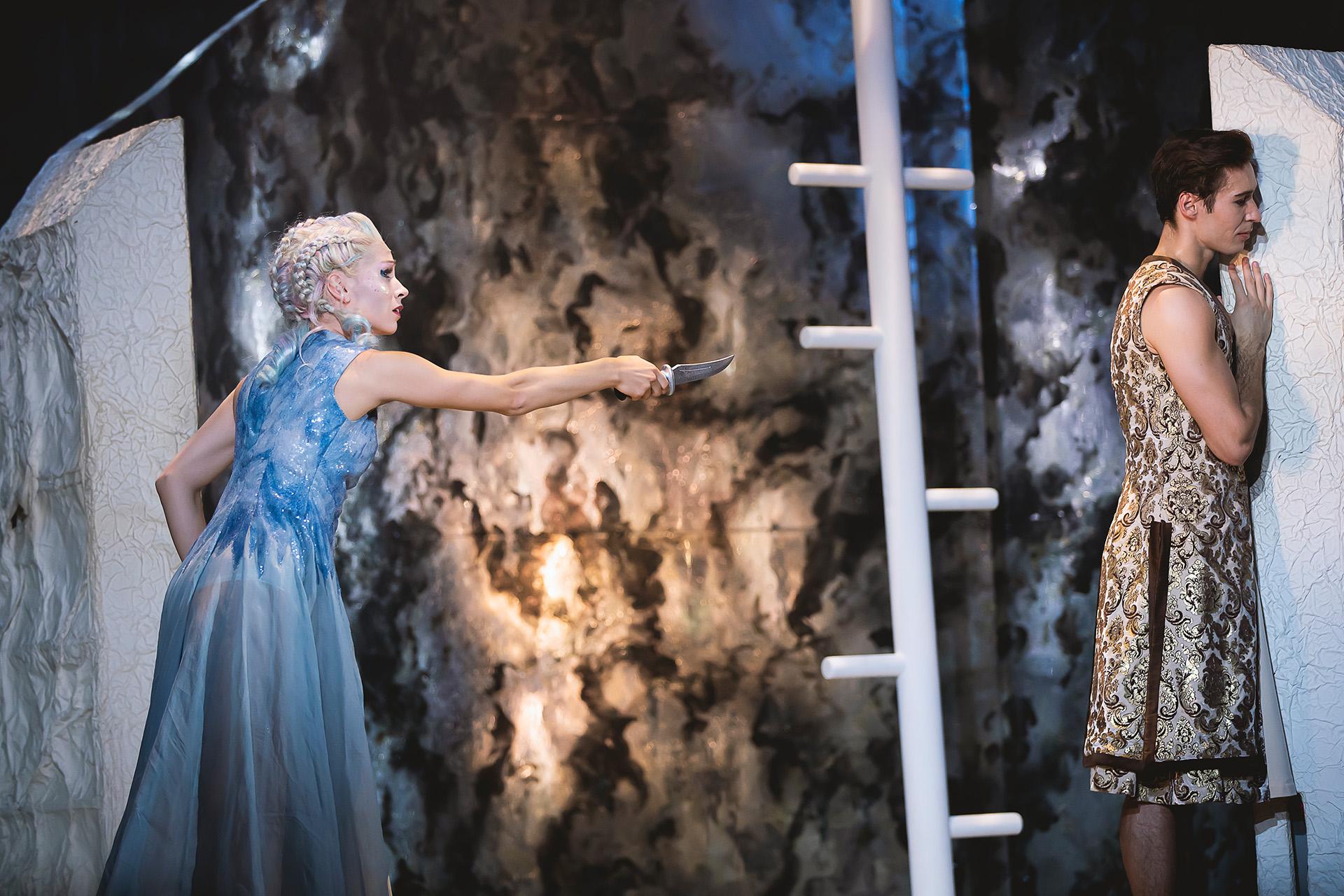 Marilla holds a knife quizzically while Prince Adair stands facing away from her