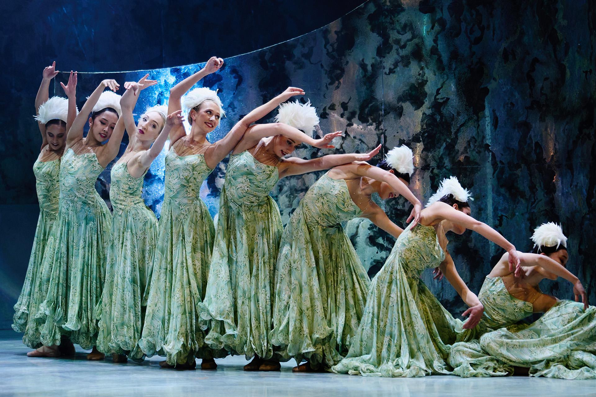 Female dancers wearing green dresses and white hats reaching up and gradually kneeling down.