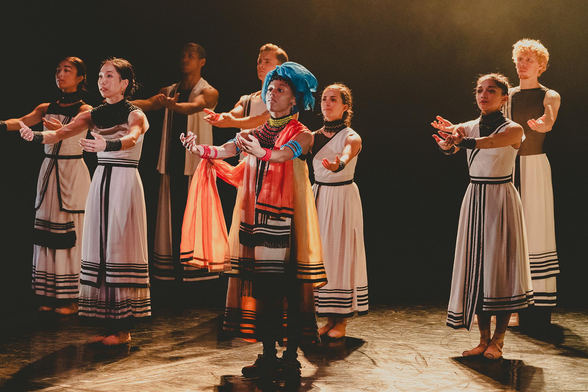 Group of dancers stand, their heads held high and their arms outstretched in front of them