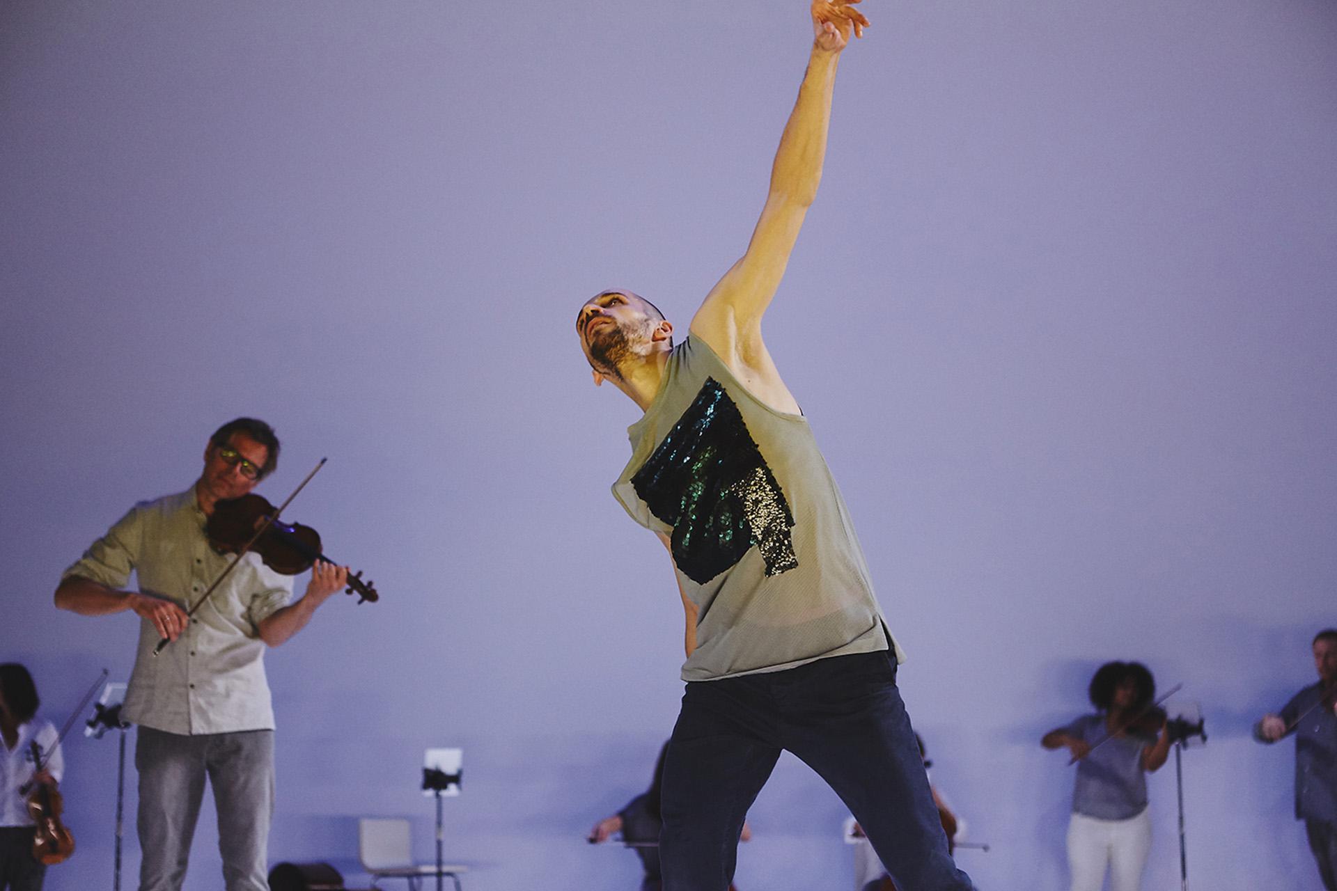 One man with a beard raisines one arm above him head triumphantly, in the background a second man in glasses and a white shirt plays the violin