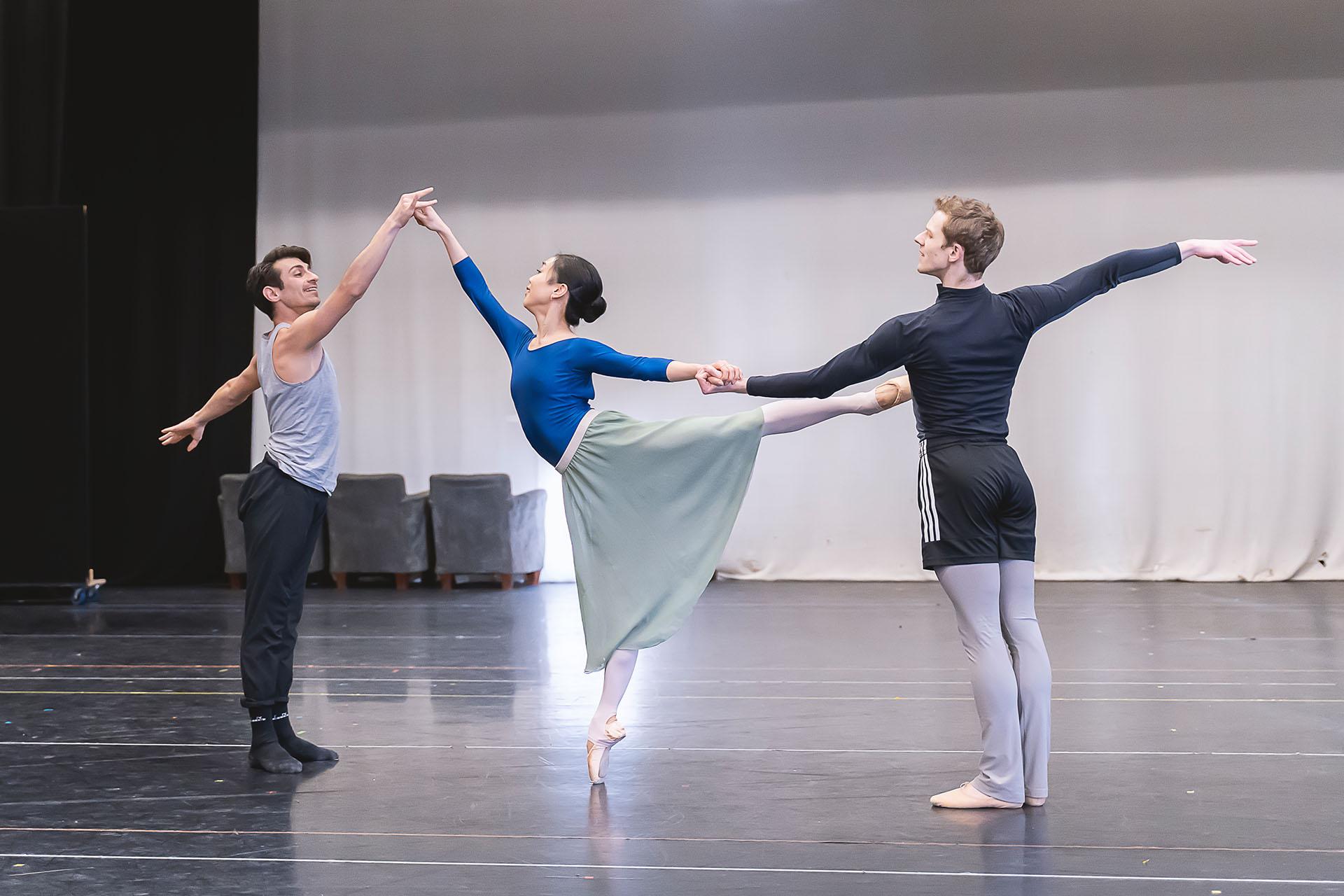 Three dancers in rehearsal, the woman dancer standing arabesque en pointe supported by one dancer behind her and one holding her hand above her head in front