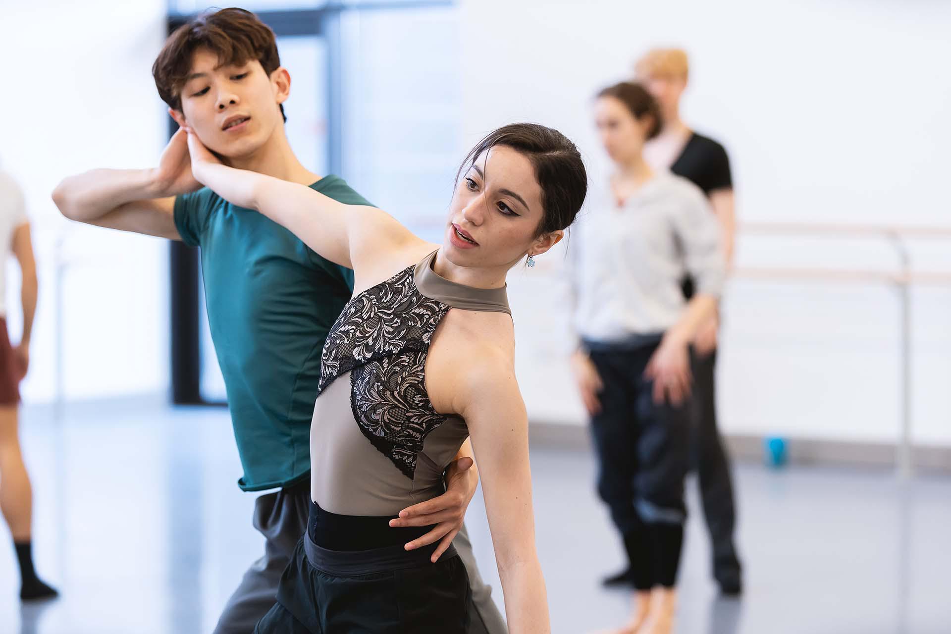 Dancer in a green top hold a hand against another's hips as she reaches bach to brush her hand against his face.