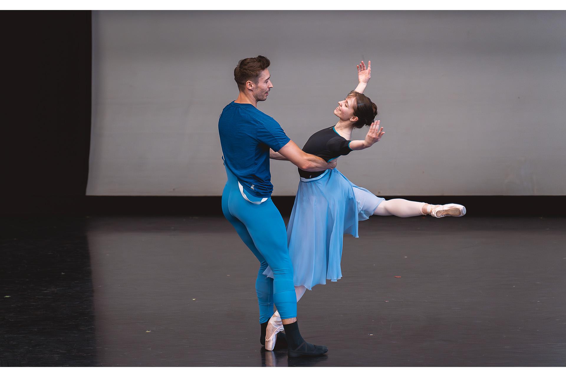 Two dancers, one supporting the other around the waist as she stands en pointe on one foot, her leg raid behind her, her back slightly arched and her arms raised to the side