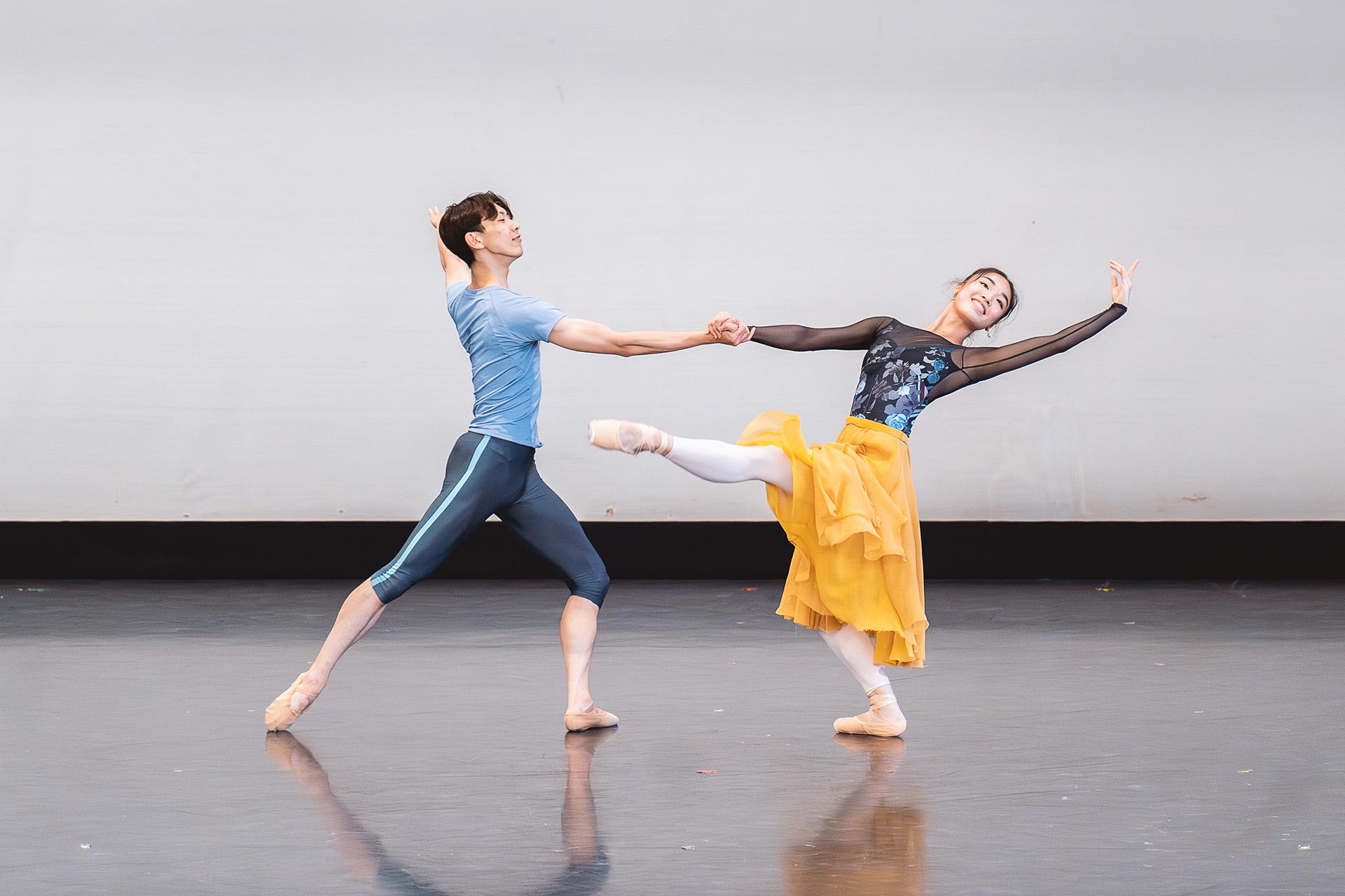 Two smiling dancers, one supporting the other with an outstretched hand, and the other, in a yellow dress and dark patterned top, stands on one leg, her other stretched out in front of her