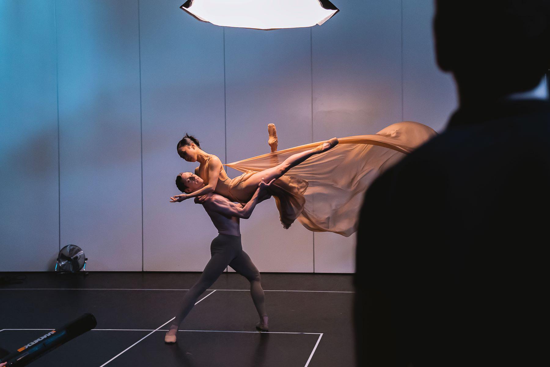 A male dancer holds a female dancer above her head in flowing fabrics.