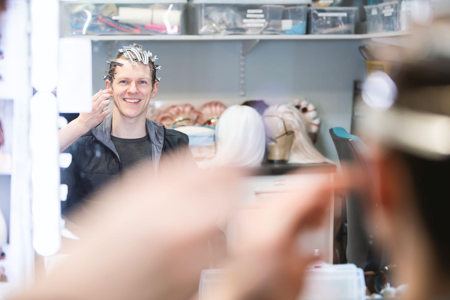 A male dancer looks at his reflection in the mirror as a stylist treats his hair.