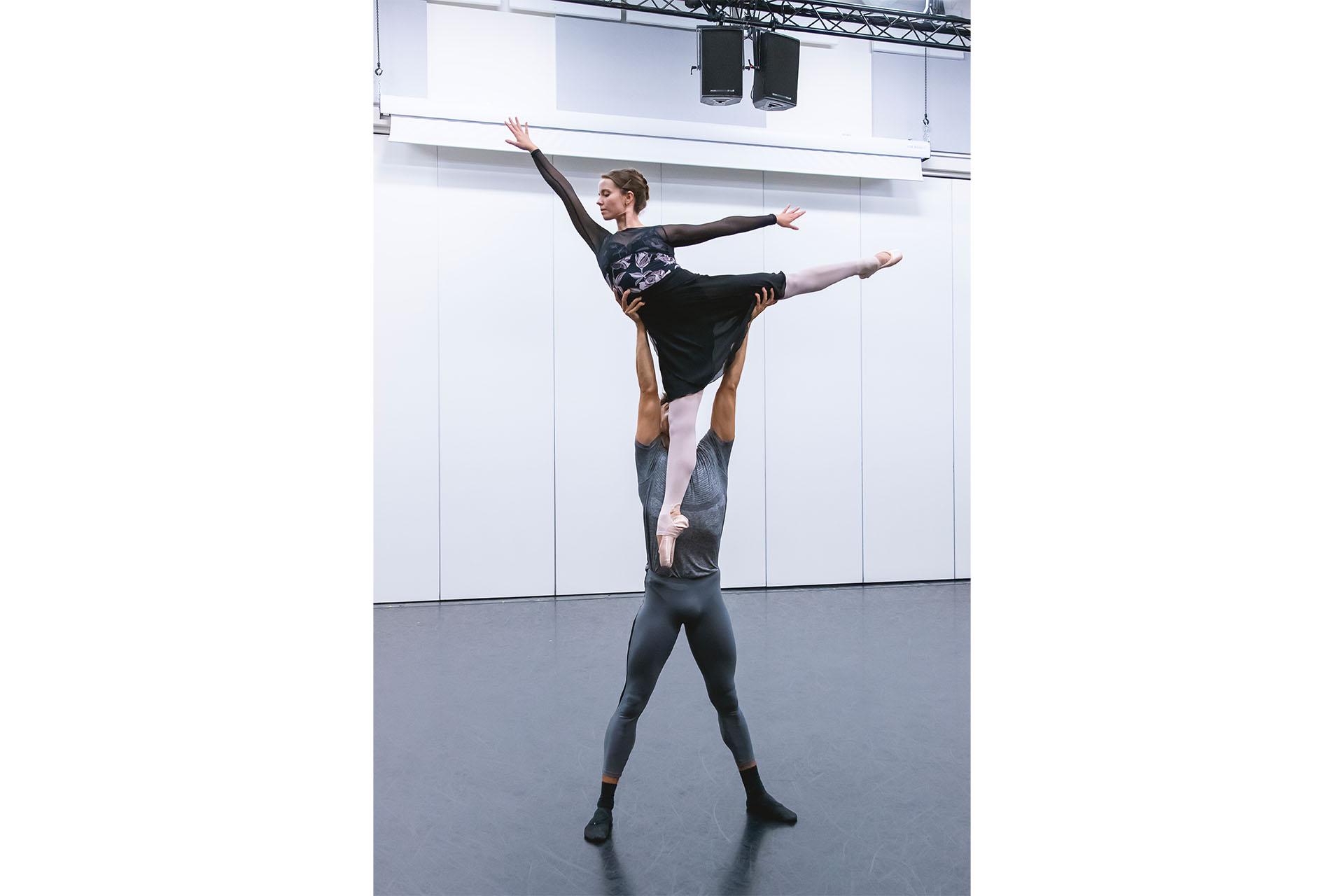 Male dancer lifts female dancer above his head, his arms fully extended, one hand by her waist and another holding her leg as she is in a classic arabesque pose.