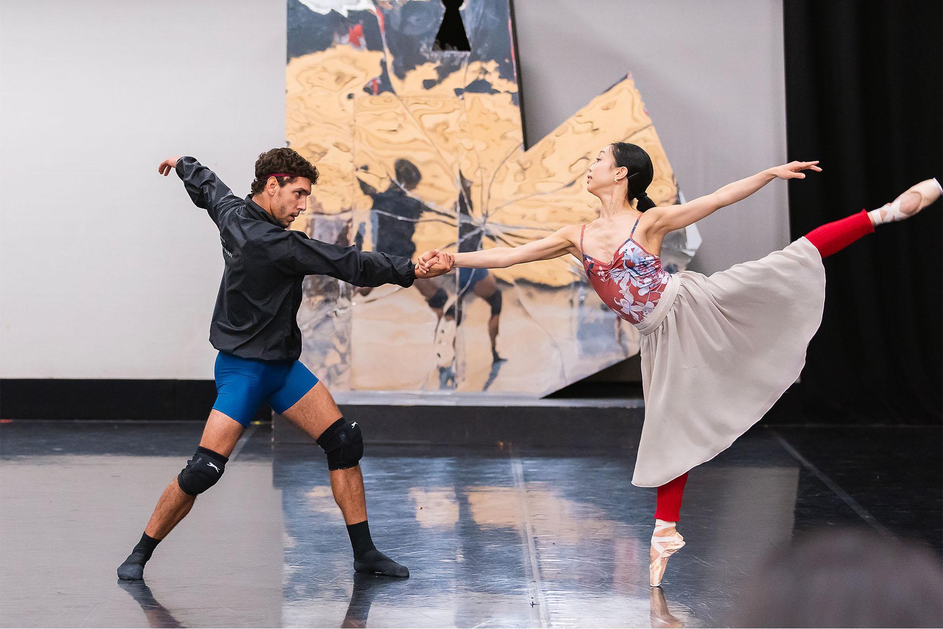 Sarah Chun in rehearsals stands en pointe on one foot, her other leg raise behind her, one arm reaching out in front of her where she holds on to a male dancer supporting her position and holding her steady.