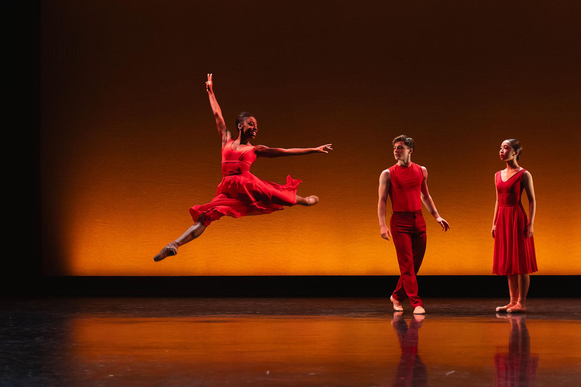 Female dancer wearing a red dress leap high with an arm stretched above her while two dancers dressed in red look on
