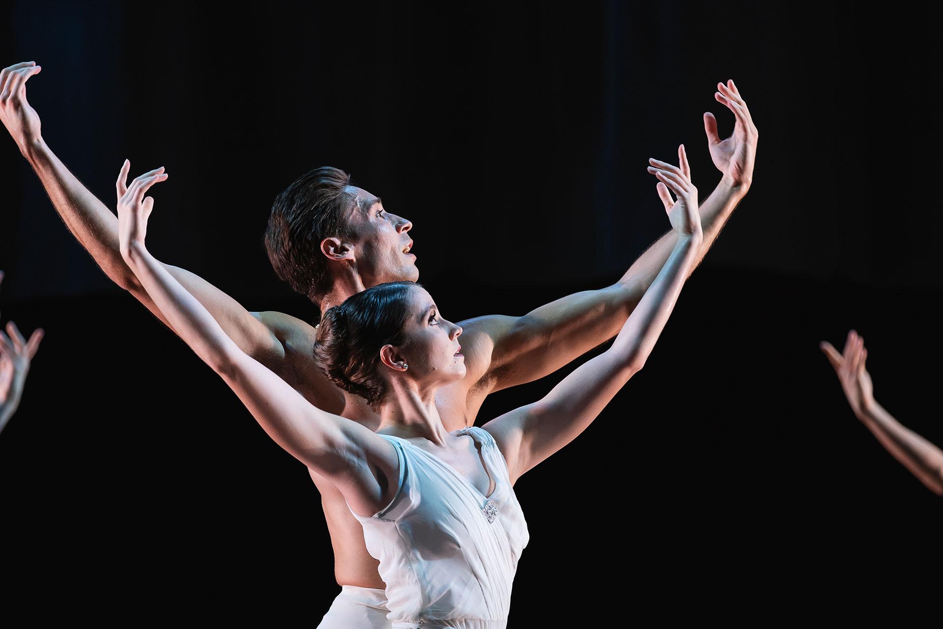 Dancers in white standing one behind the other, half look to the sky with their arms raise like Atlas