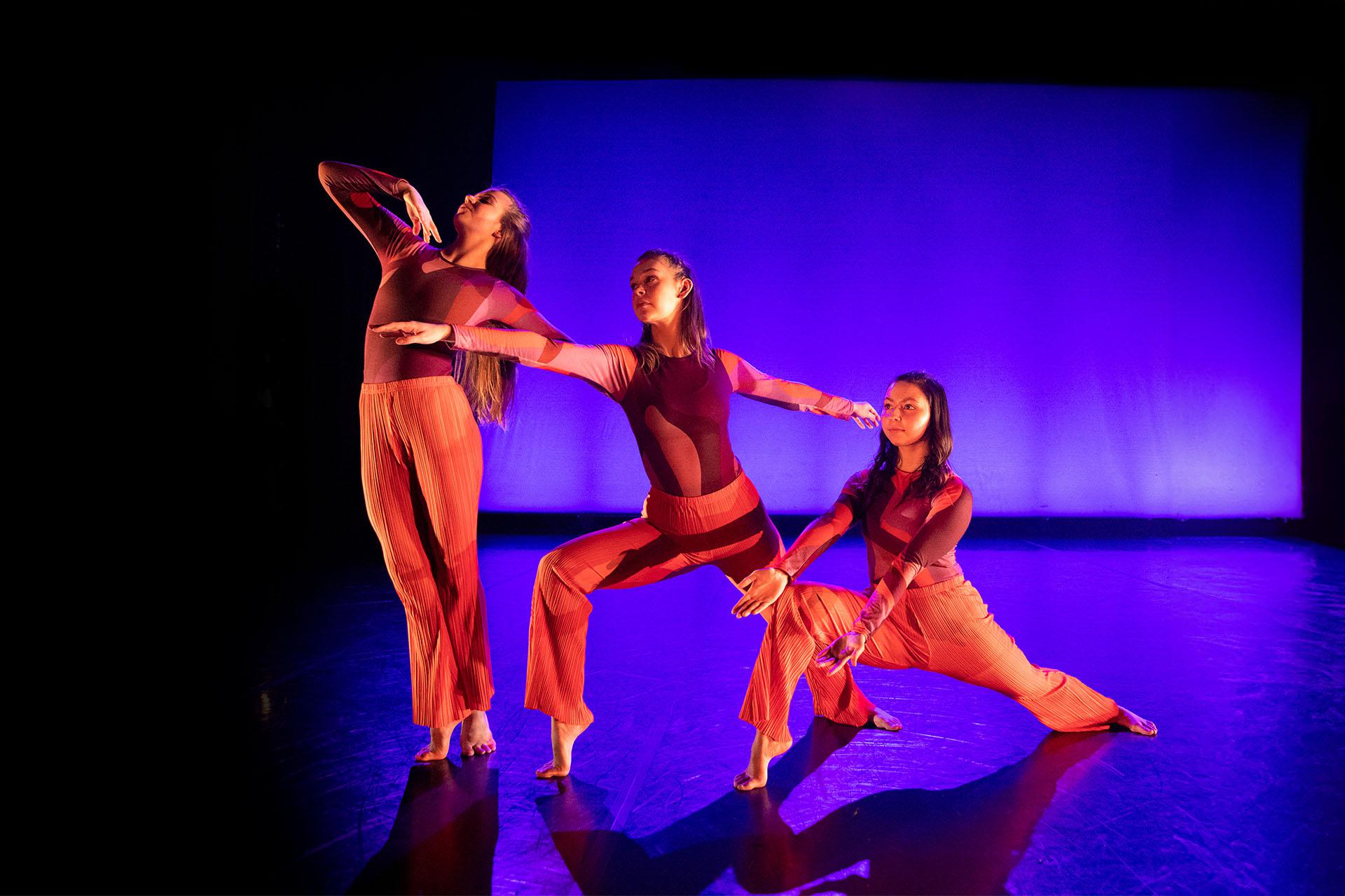 Three dancers bathed in red light against a background of black and blue, each in a different pose - one on her toes sood tall, the next bending one leg at 90 degrees, the third lower still