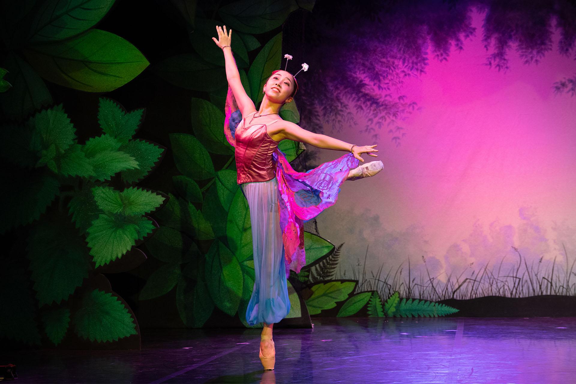 Female dancer, dressed as a butterfly in magenta and cyan, stands arabesque en pointe