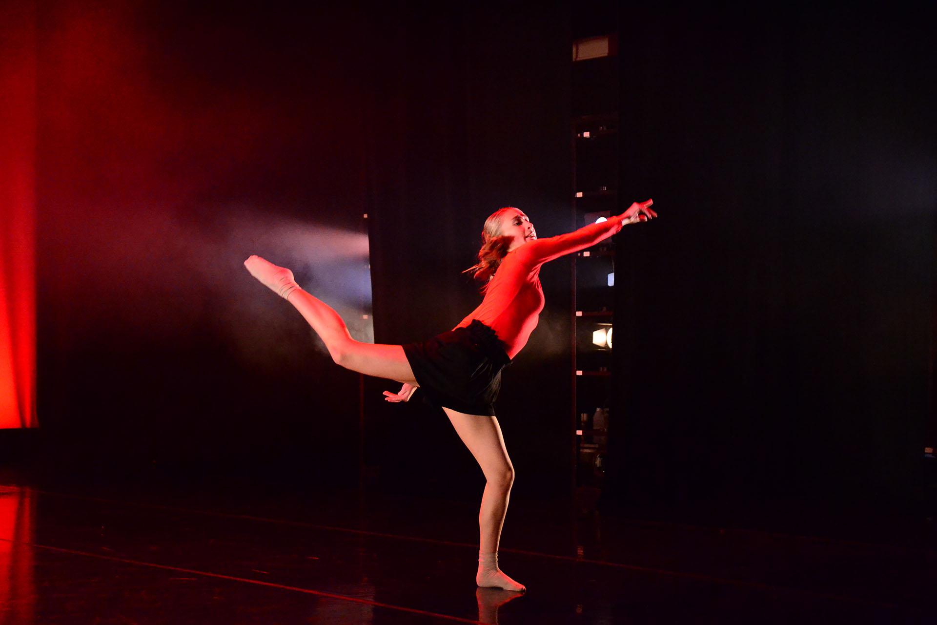 Against a dark background a female dancer stands on one foot, the other leg raised behind her and her body leaning forward and pointing