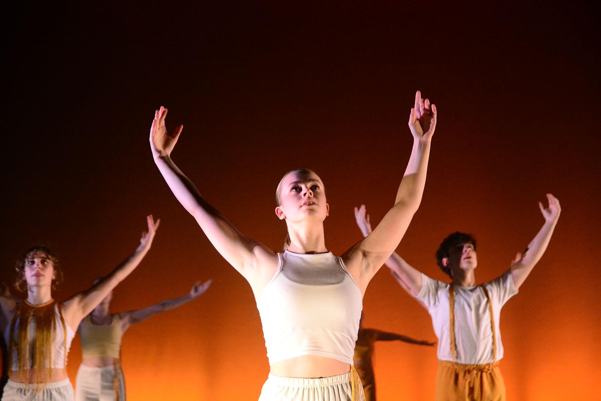 Young female dancer in a white top against a lit orange background looks into the air and raises her hands above her head