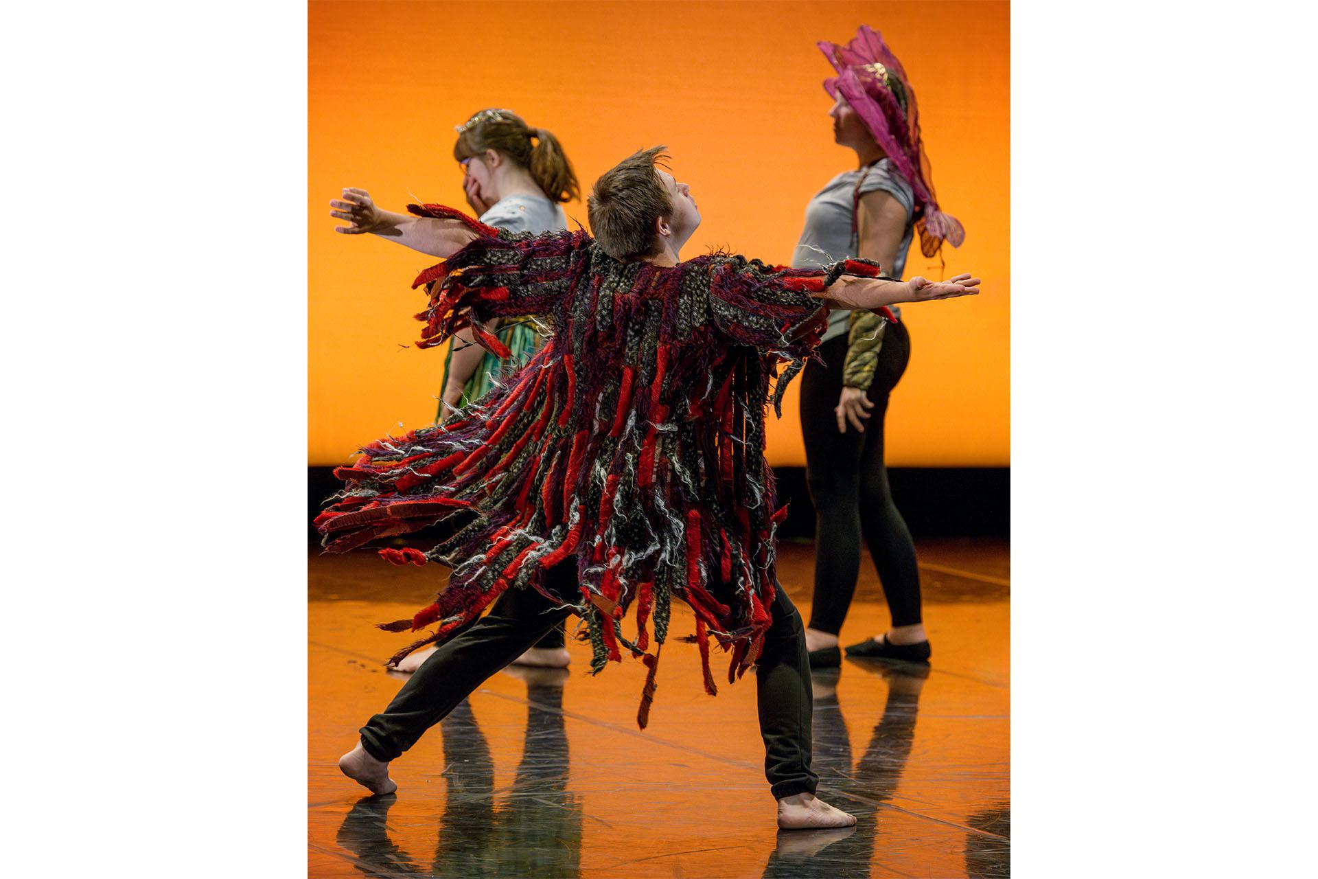 In front of an orange backdrop, a man wearing a coat made of red and black ribbons spreads his arms expressively
