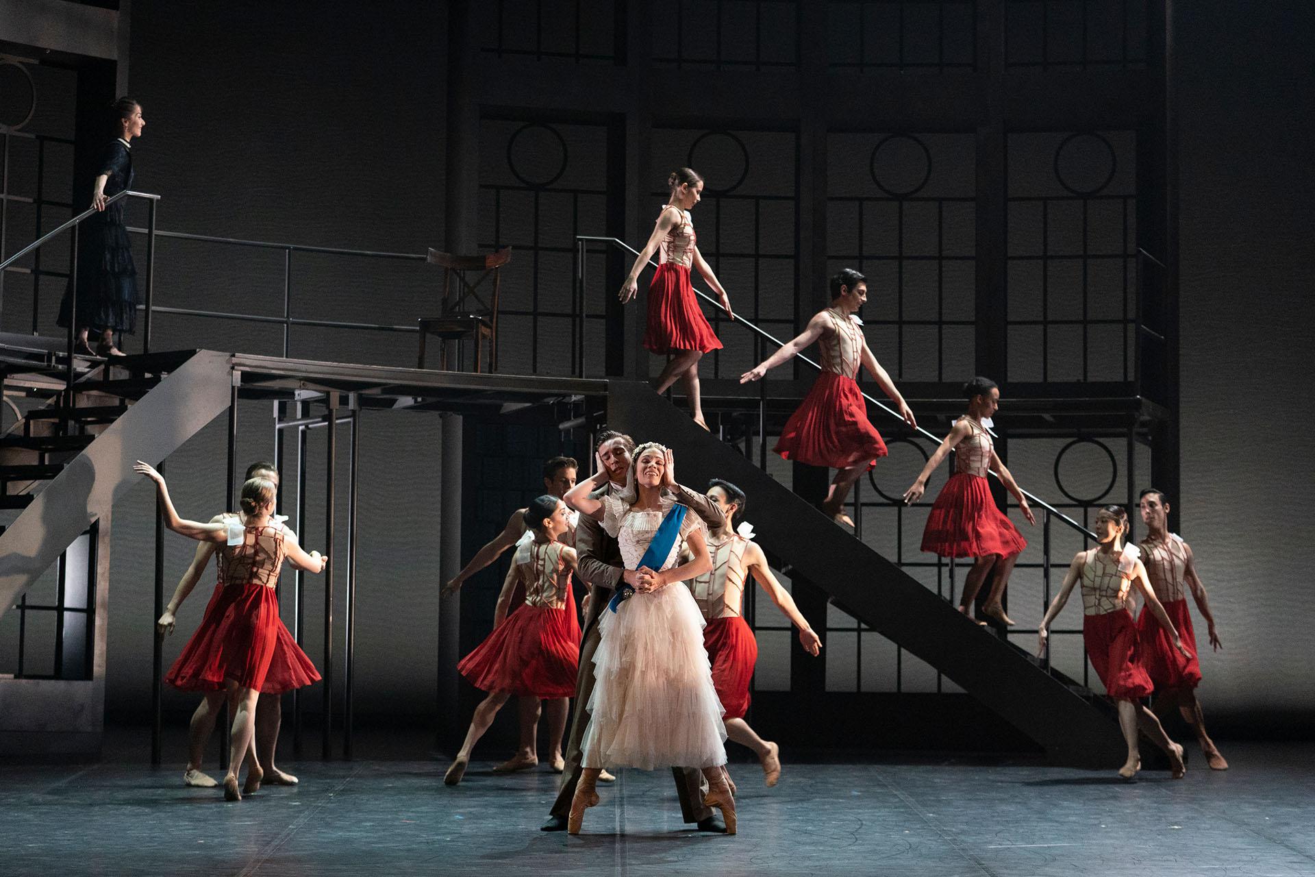 Queen Victoria in a white dress and blue sash is embraced from behind by her husband white dancers in red and white are in the background moving and organising