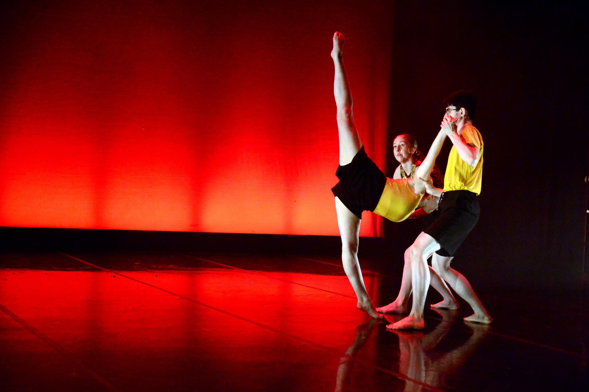 Three dancers in black shorts and yellow vests, one leaning forward with one leg on the ground and the other 180 degrees, the two remaining dancers are holding her arms supporting her