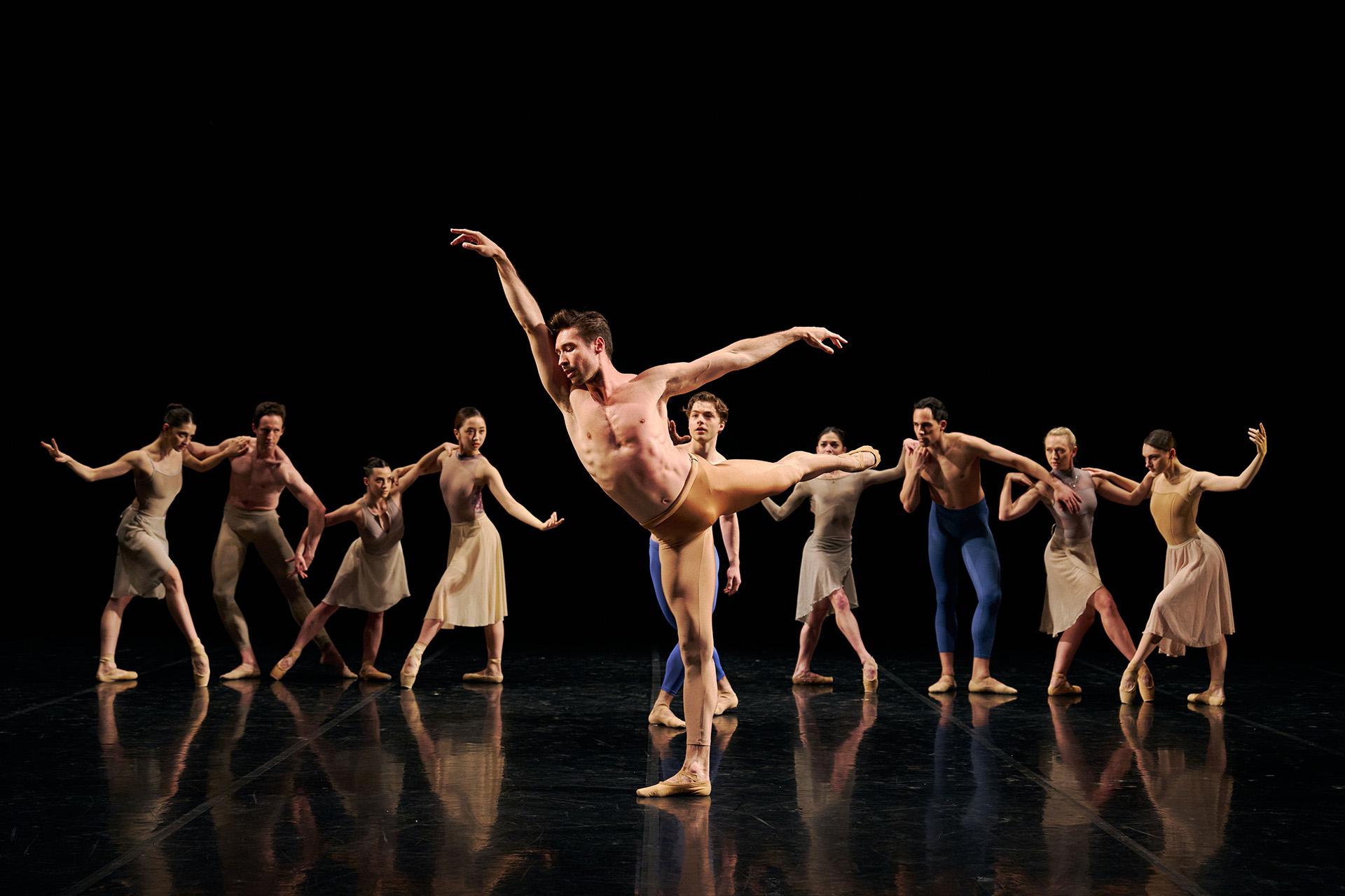 Dancer wearing gold shorts and nothing else stands arabesque while nine other dancers pose behind him watching
