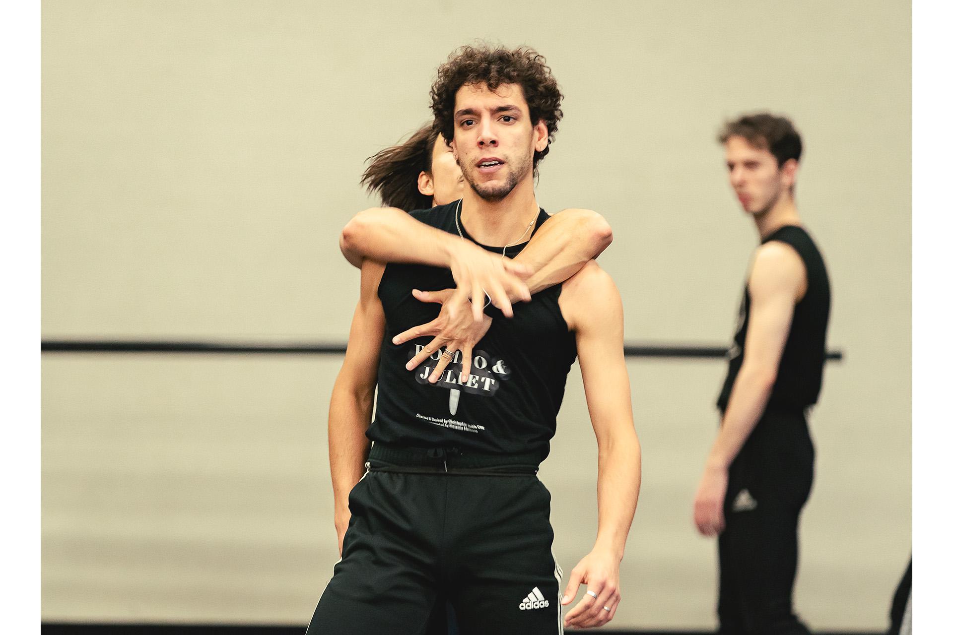 Dancer with short dark hair stands with another arms coming from behind around him