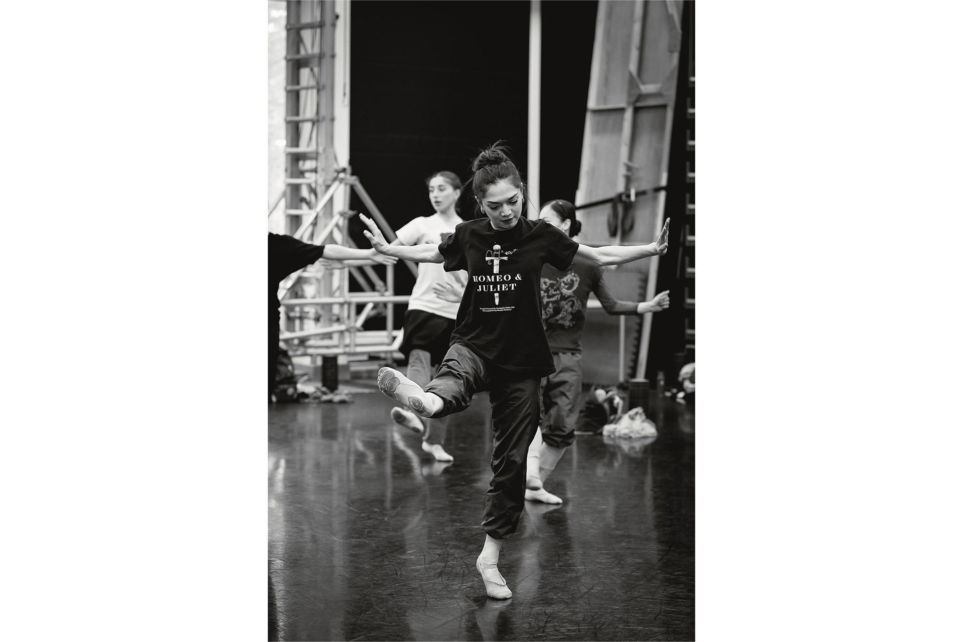Black &amp; white image, in rehearsal a dancer stands on one foot, the other extended to the front, her arms wide to the sides