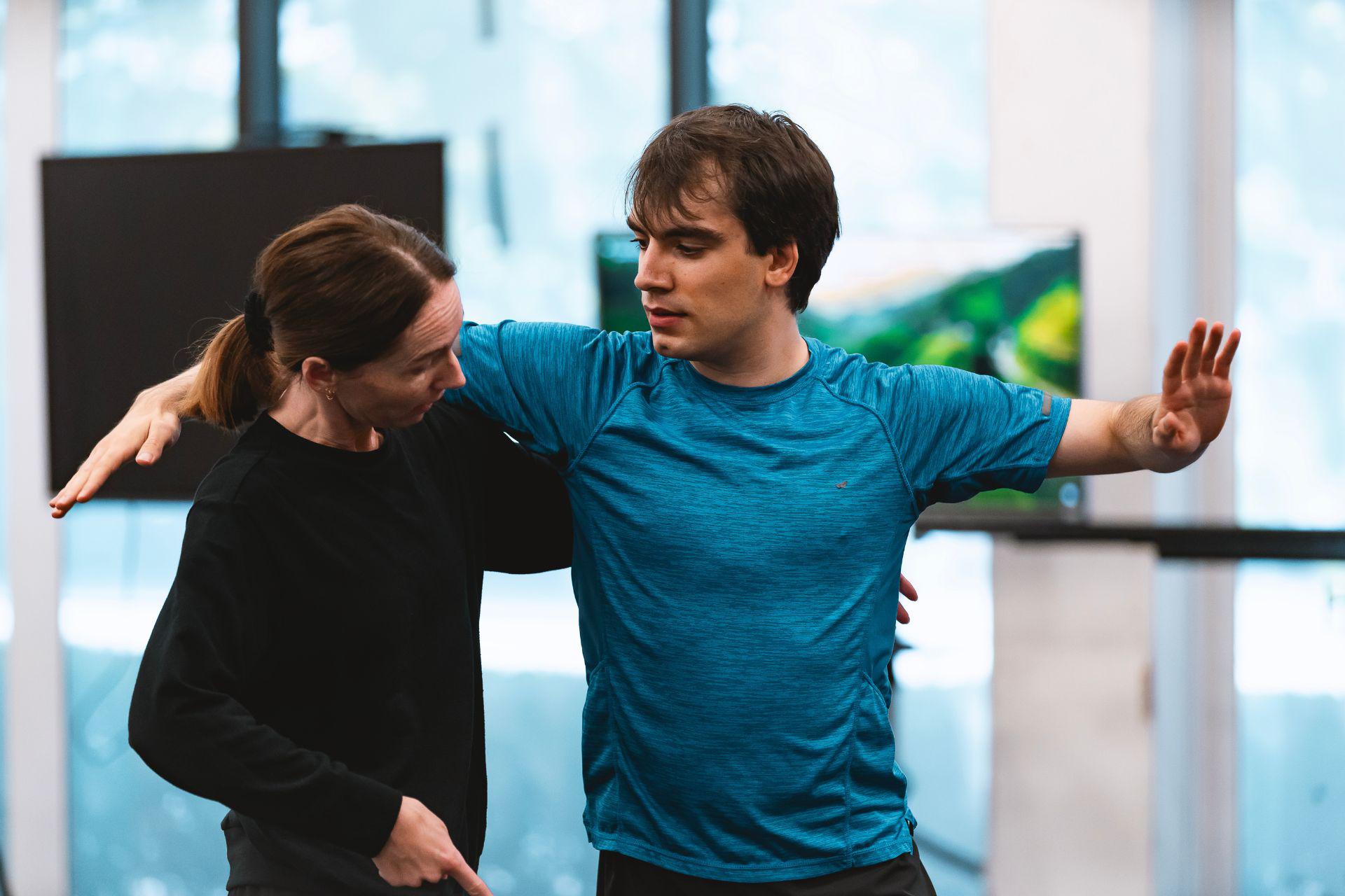 A choreographer has her hand on a dancers waist. The dancer watches her, his arms raised at his side.