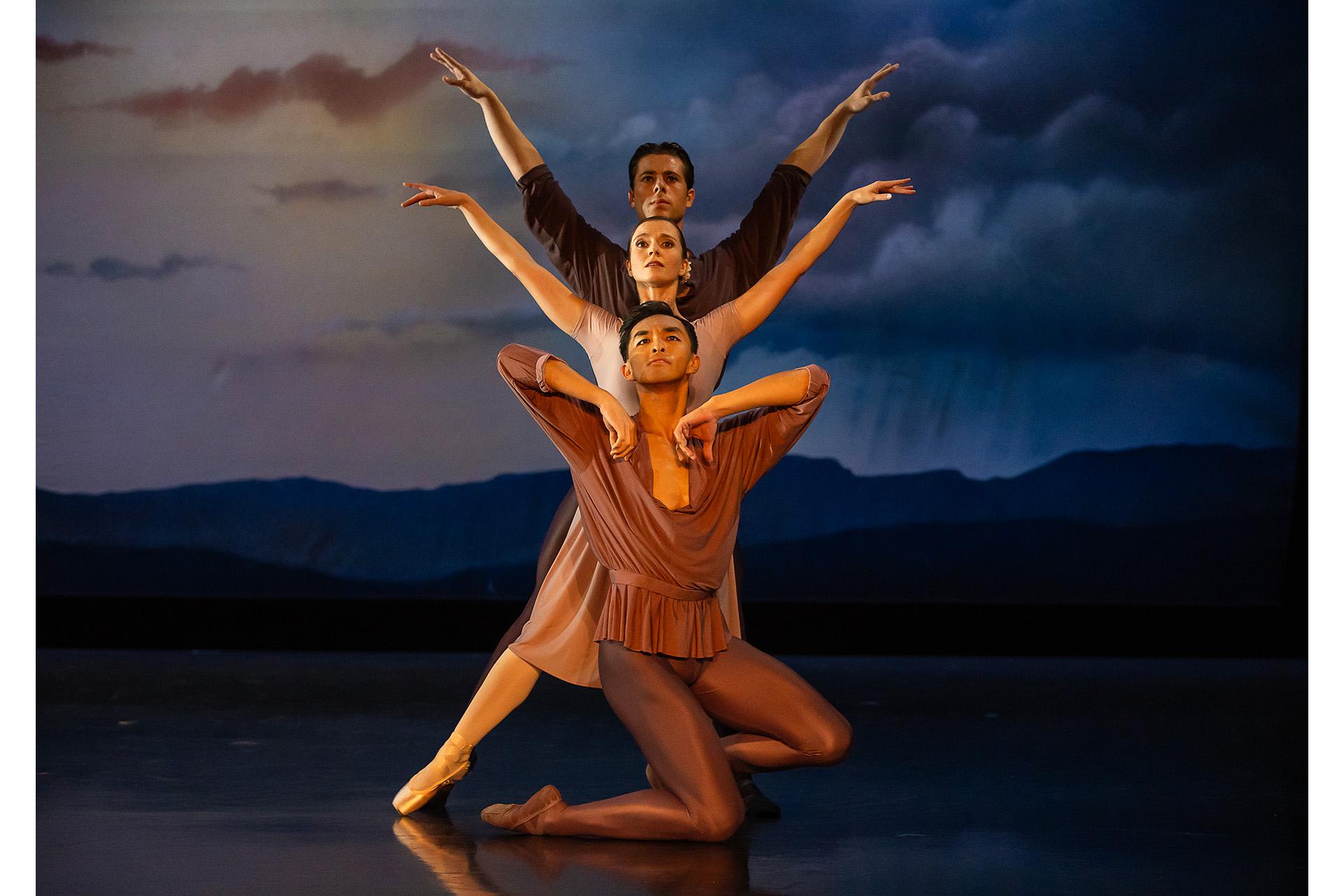 Three dancers pose immediate behind one another, the first dressing in brown kneels, behind him a dancer stands with her arms raised, similarly the dancer behind her but he is taller