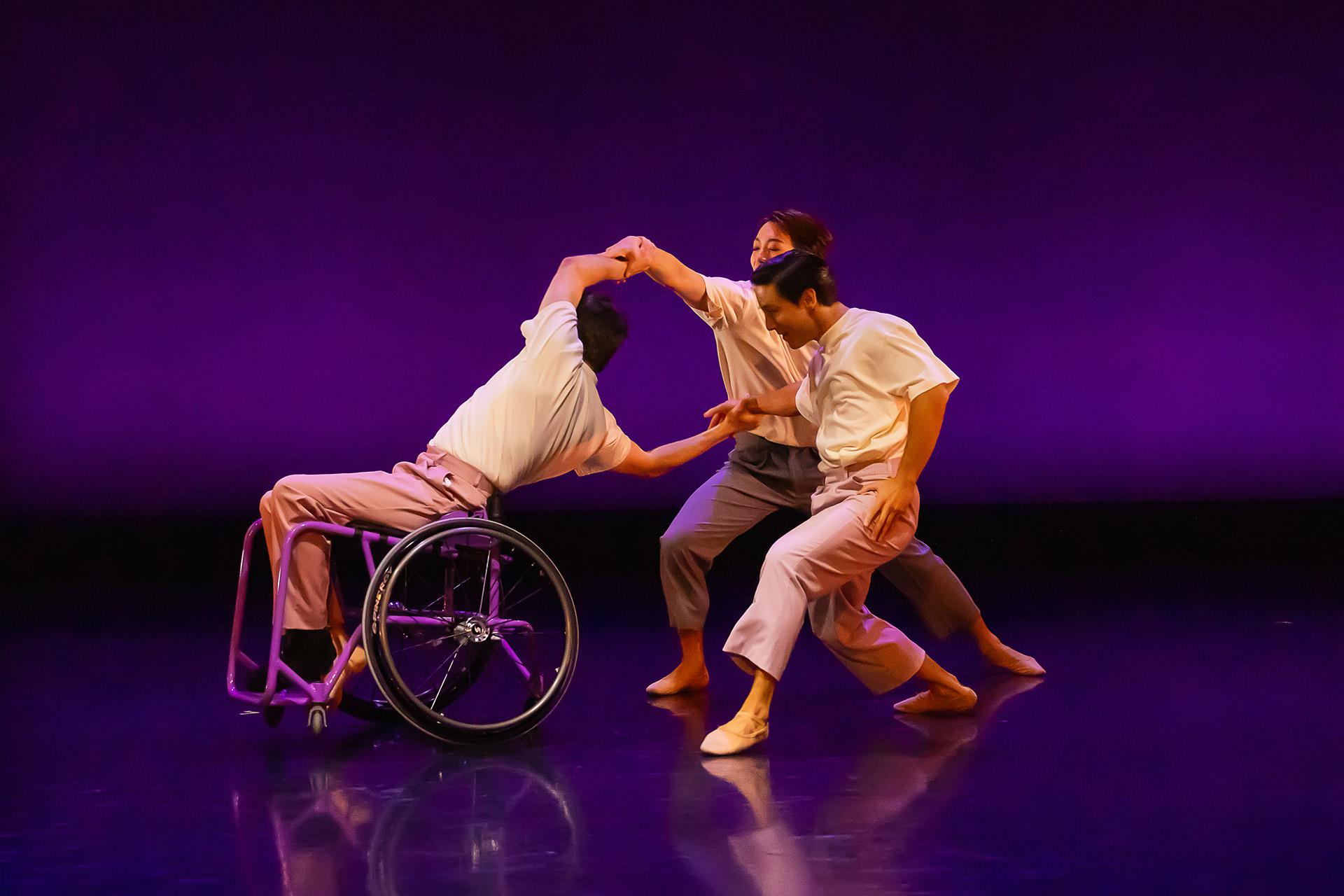 One dancer in a wheelchair leans back and holds the arms of two more dancers immediately behind him