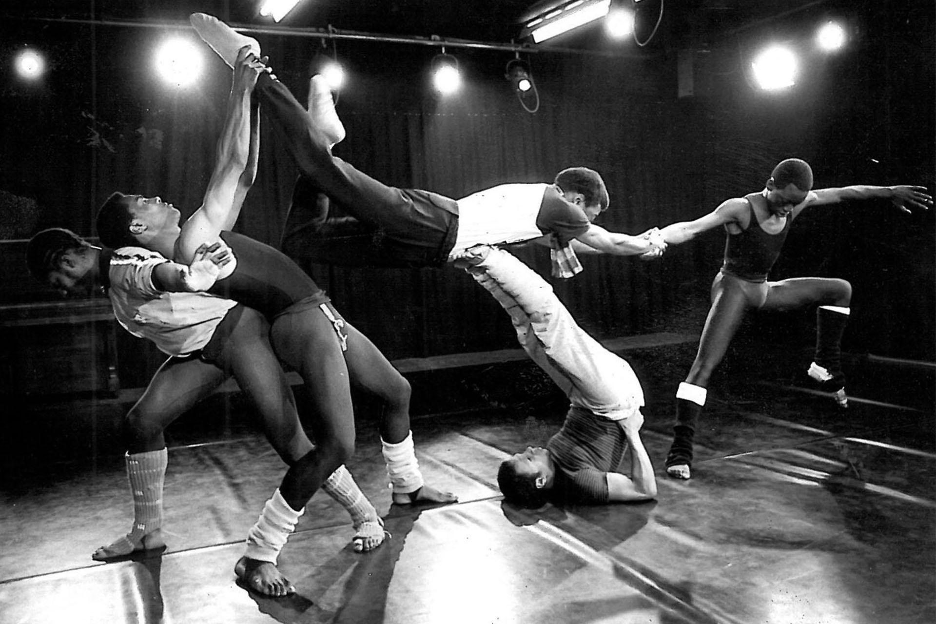 Monochrome image of five black people dancing, supporting each other, one lifting another while laying down, another leaning, one providing balance by holding and pulling