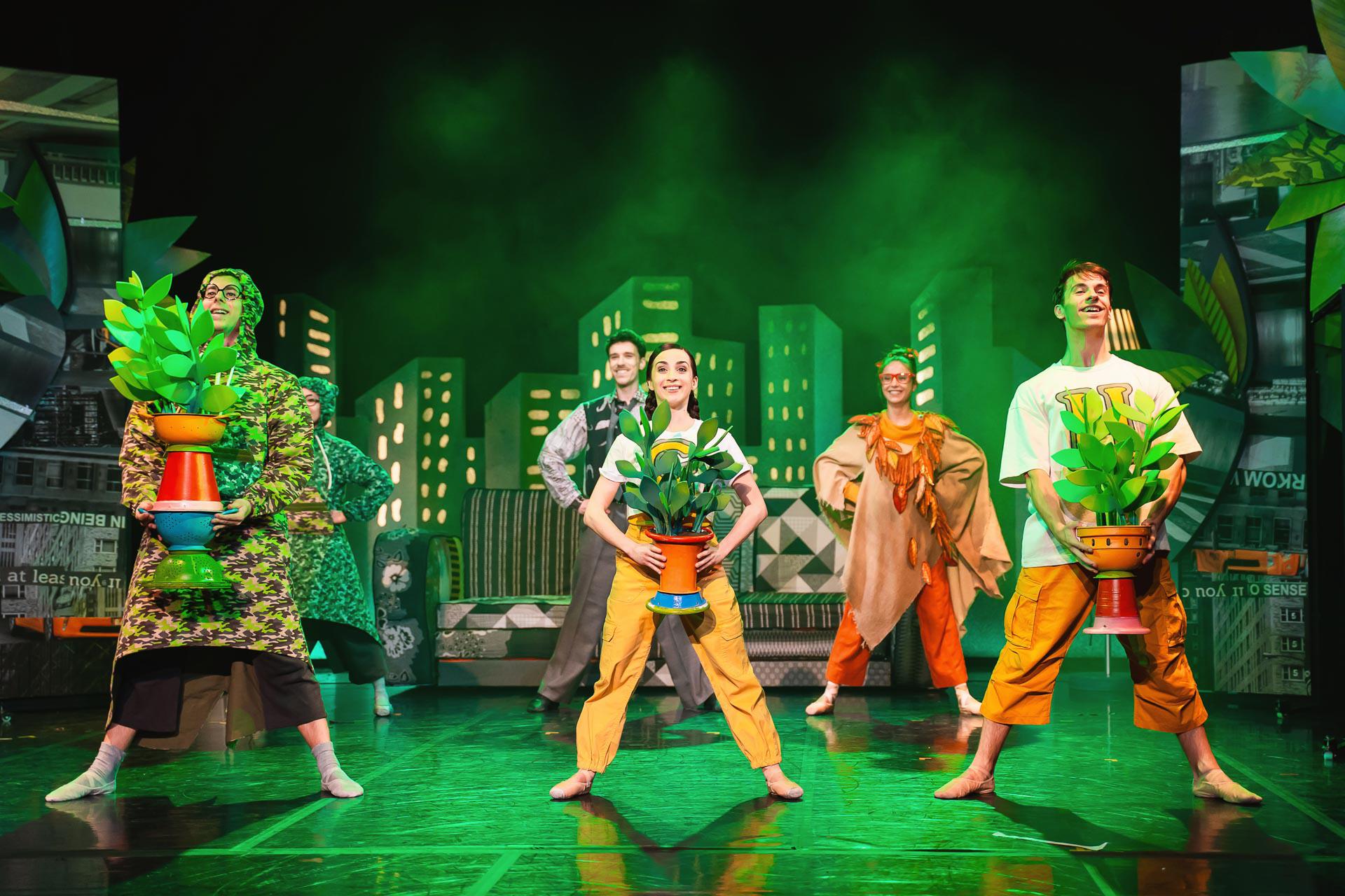 Dancers stood in a row holding potted plants in a soft green light