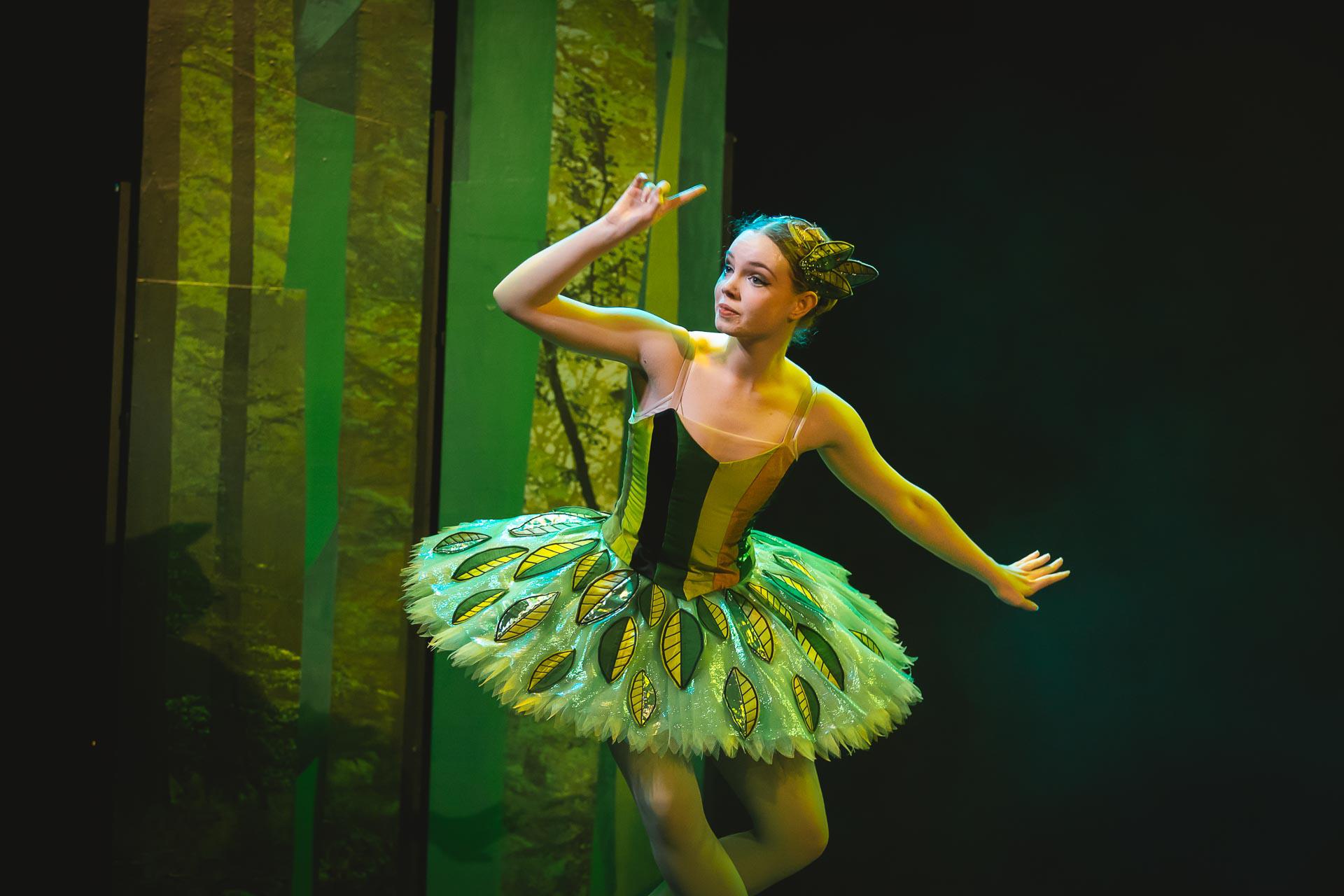 A dancer stood in a green tutu with one arm raised with a finger extended to her forehead. Her costume is covered in green leaves