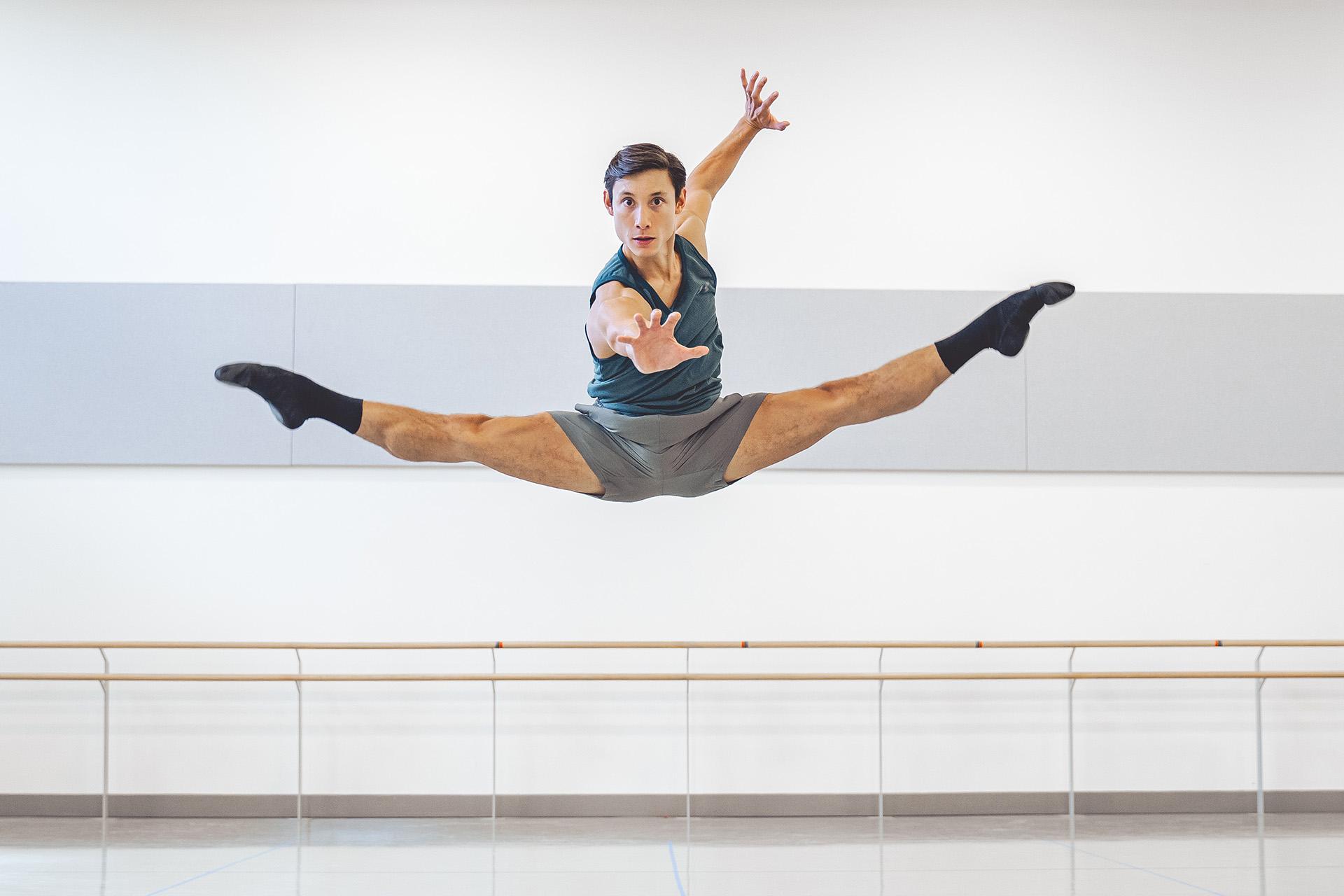 A dark haired dancer leaps, legs spread mid-jump, one hand outstretched to the camara